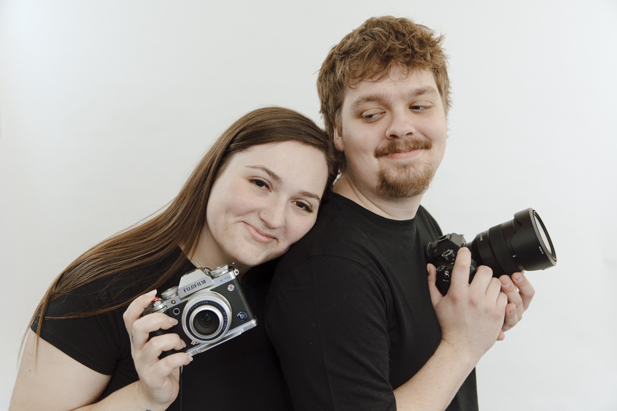 A young woman and a young man are taking a selfie together, both smiling and holding cameras, with a plain white background.