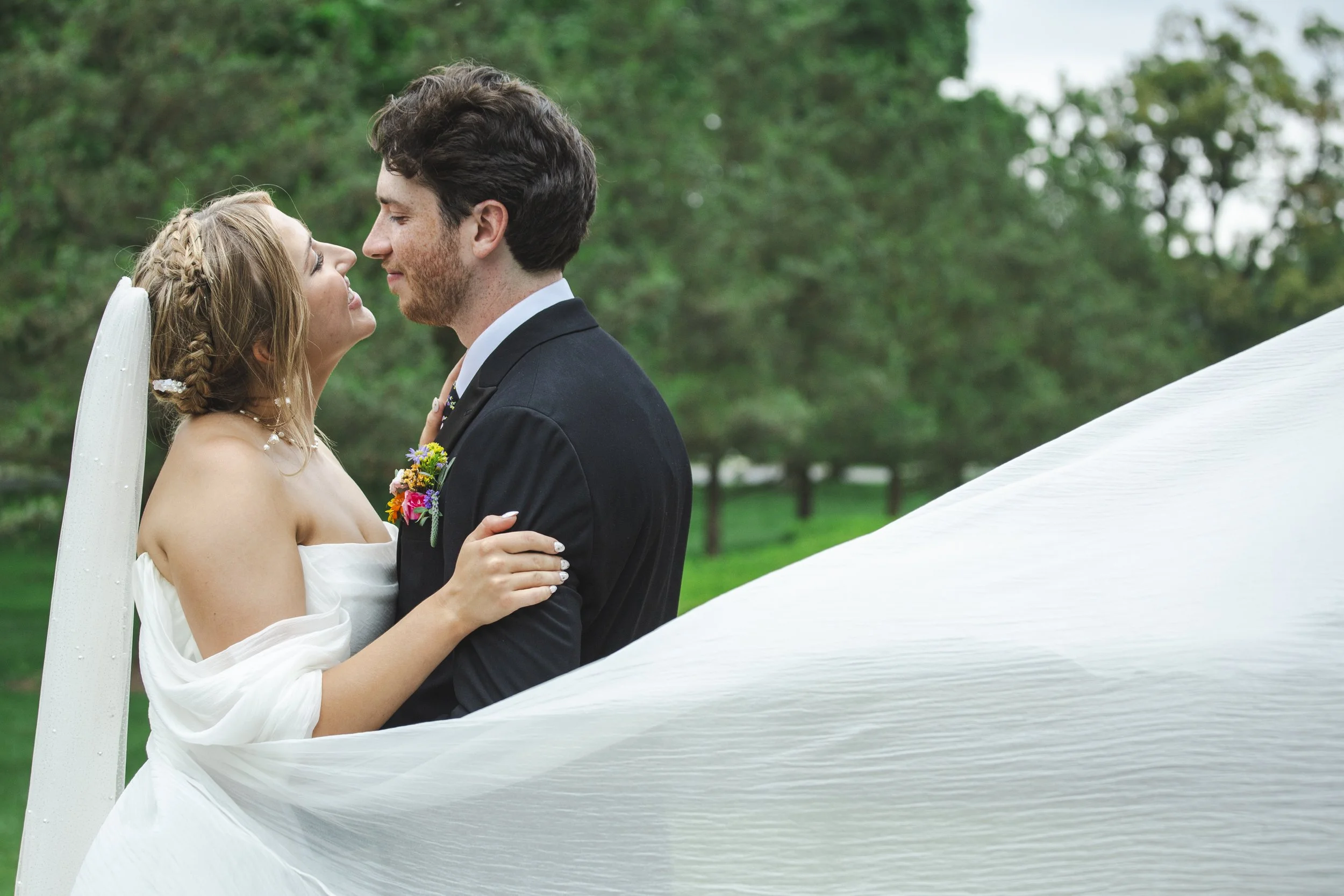 A bride and groom share a kiss outdoors on their wedding day, with green trees in the background.