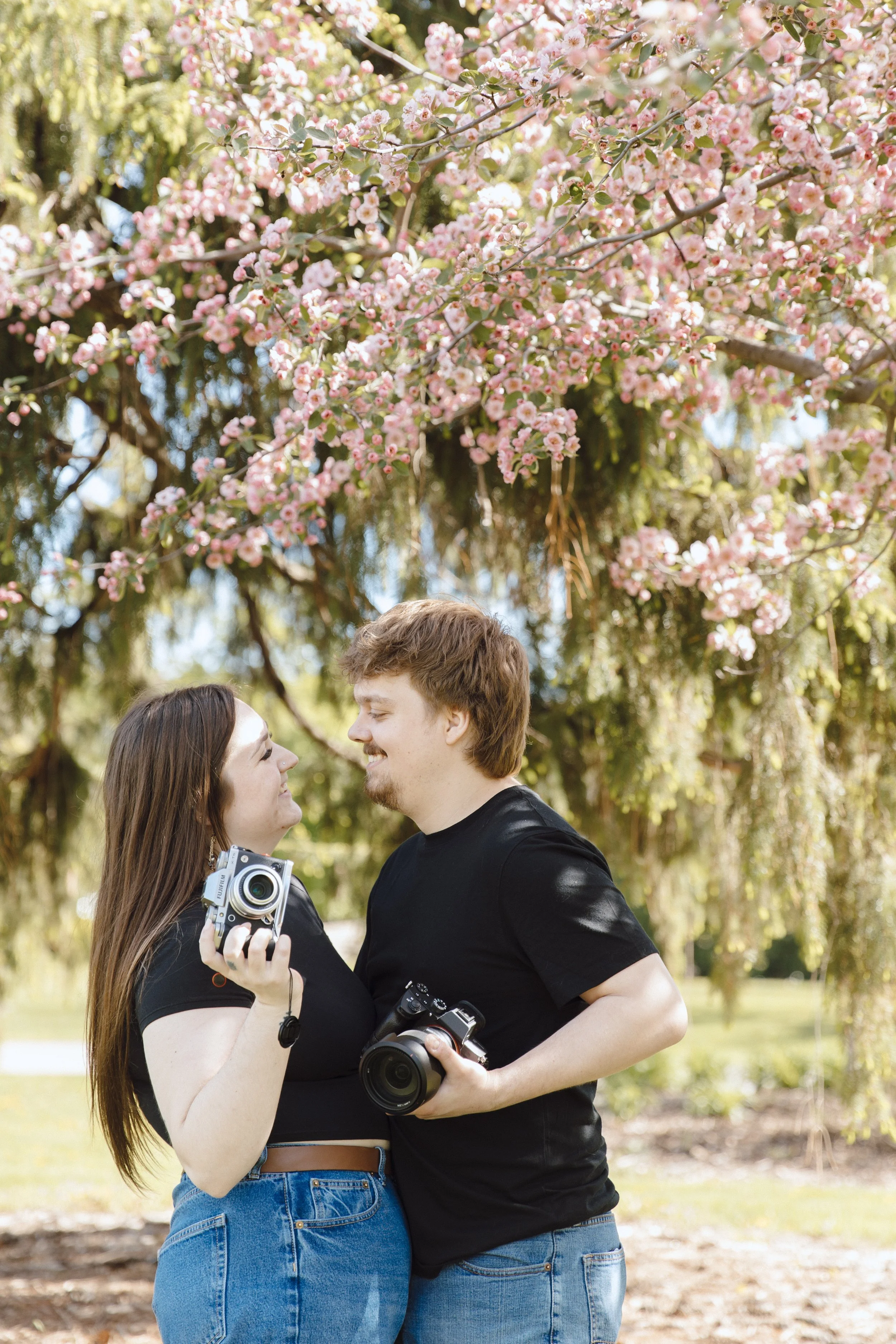 A young woman and young man stand close together outdoors under a blooming pink cherry blossom tree, holding cameras and smiling at each other.