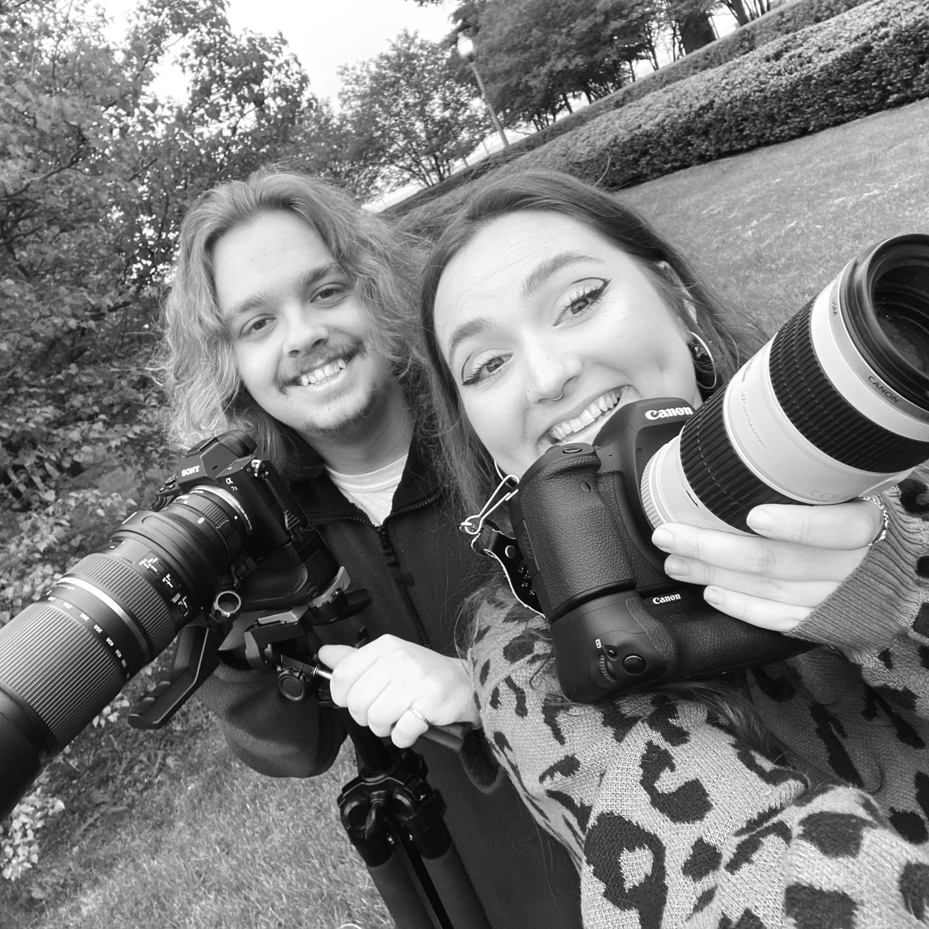 Two photographers taking a selfie outdoors in a park with trees and bushes in the background, both smiling and holding professional cameras with telephoto lenses.