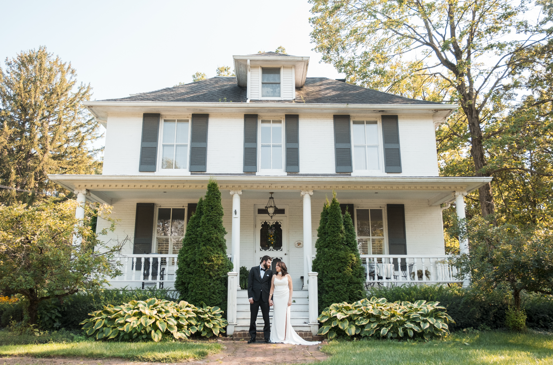 A newlywed couple dressed in wedding attire standing in front of a large white house with black shutters, two or three stories tall with a porch, surrounded by lush greenery and trees with autumn foliage.