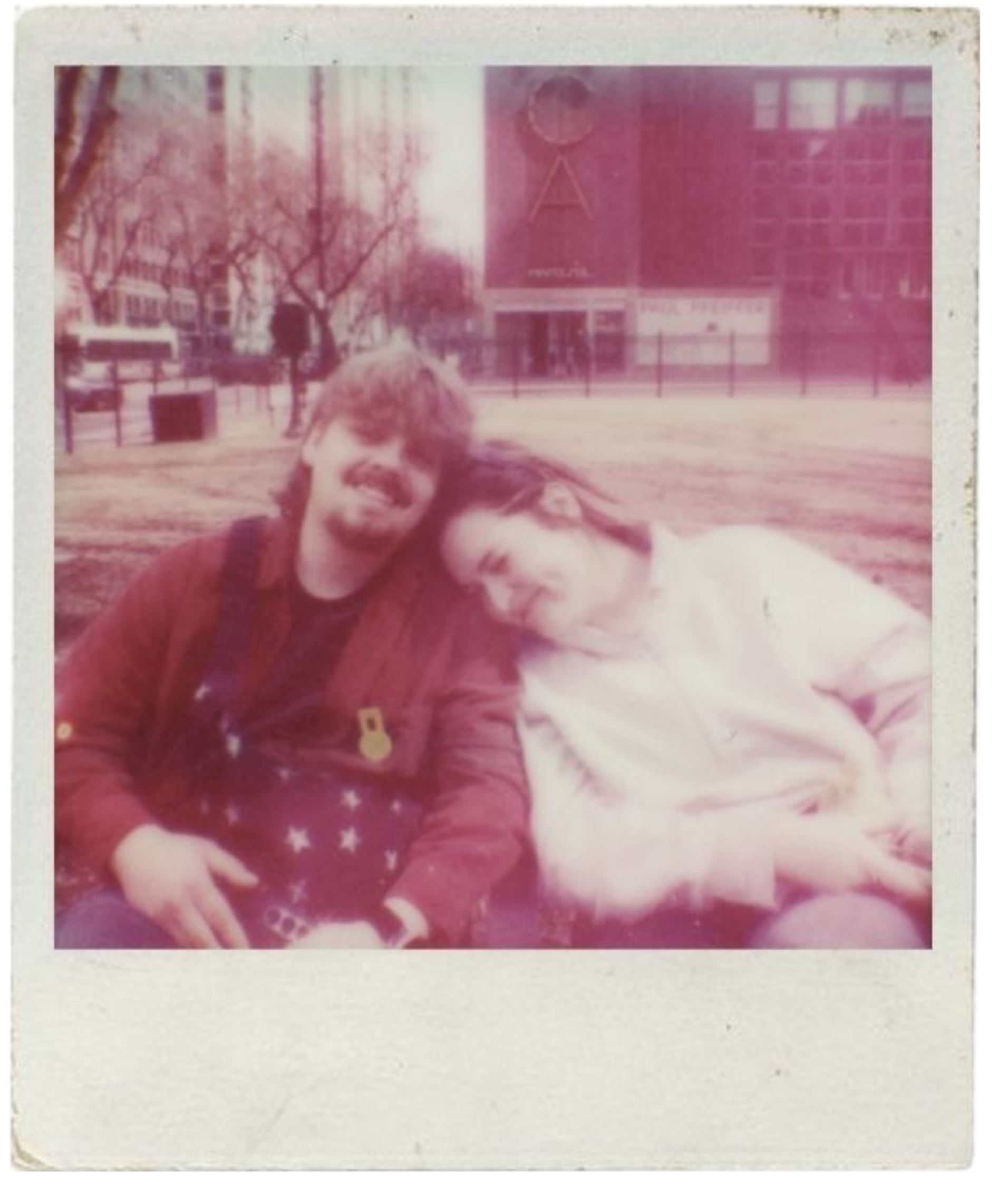 A vintage Polaroid photo of two people sitting on the grass in a park, smiling and leaning towards each other, with trees and buildings in the background.