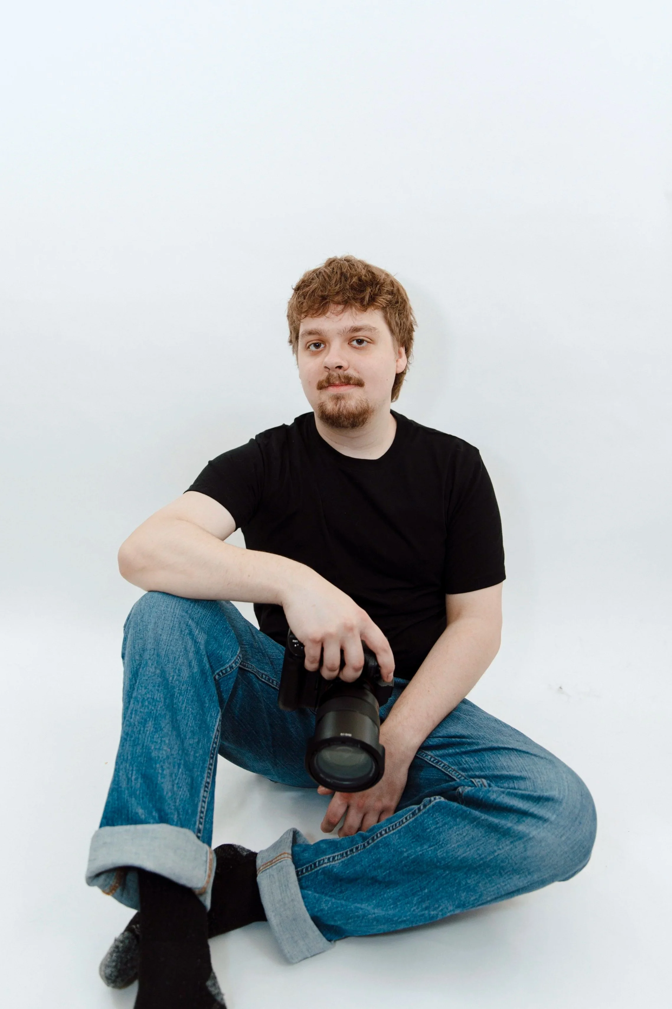 A young man with curly red hair, a beard, and mustache sitting on the floor against a plain white wall, wearing a black t-shirt, blue jeans, and black socks with gray accents, holding a camera.