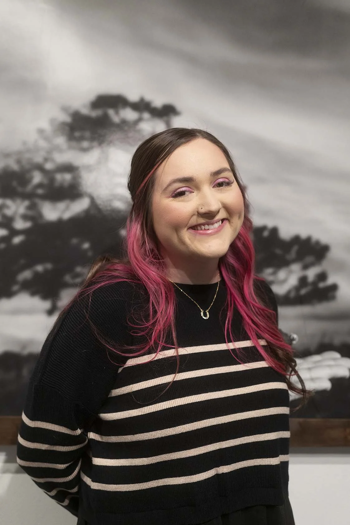 A young woman with long pink hair and a nose piercing, wearing a black and beige striped sweater, smiling in front of a black and white image of trees.