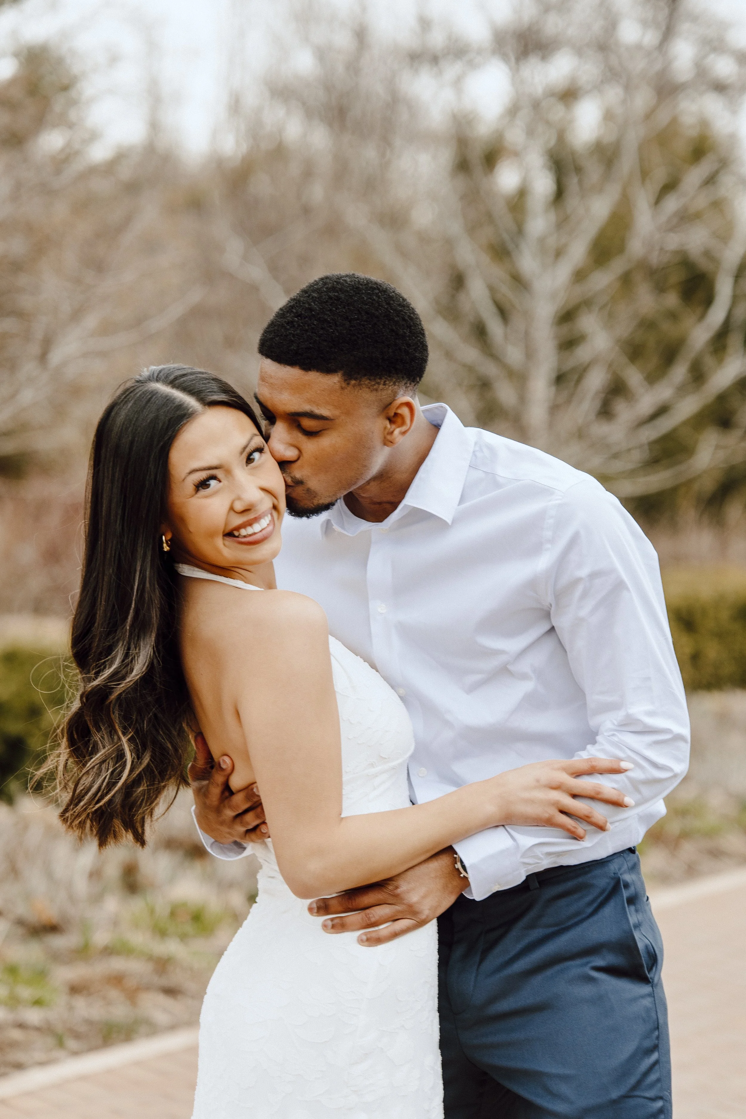 A couple hugging outdoors, the man kissing the woman's cheek, the woman smiling at the camera with long dark hair, wearing a white dress, the man wearing a white shirt and dark pants, in a natural background with trees.