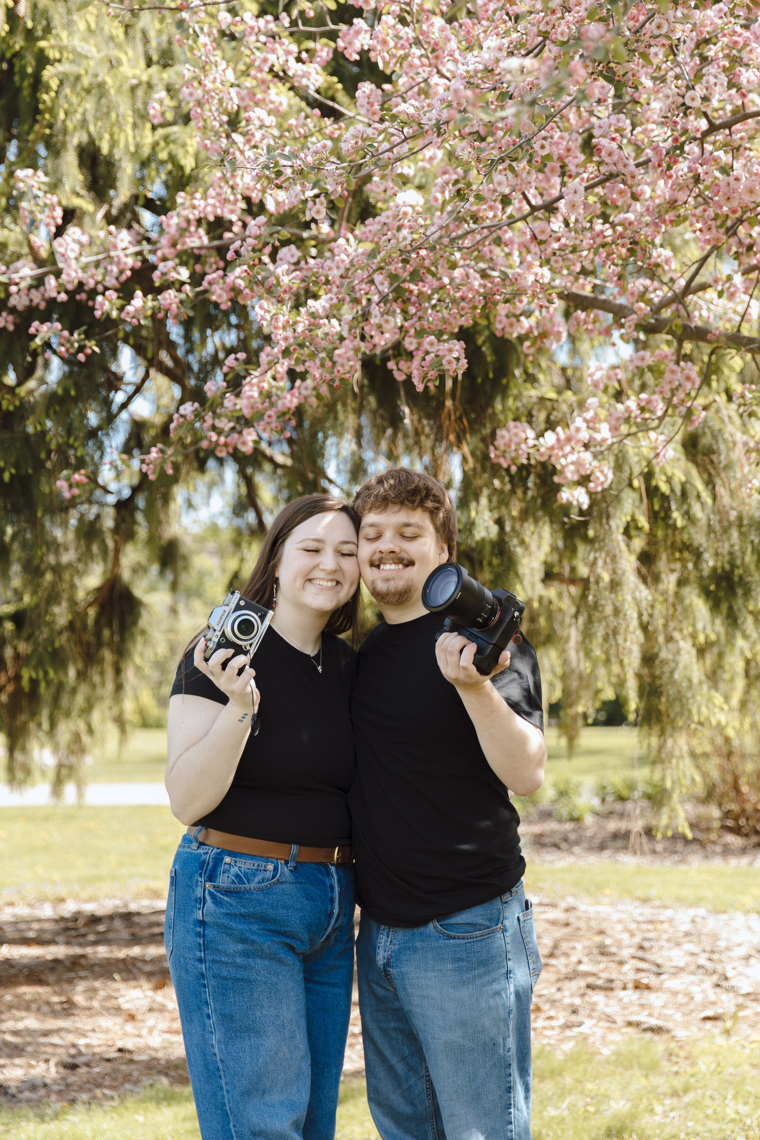A couple smiling and hugging each other outdoors under a blooming pink cherry blossom tree, holding cameras.