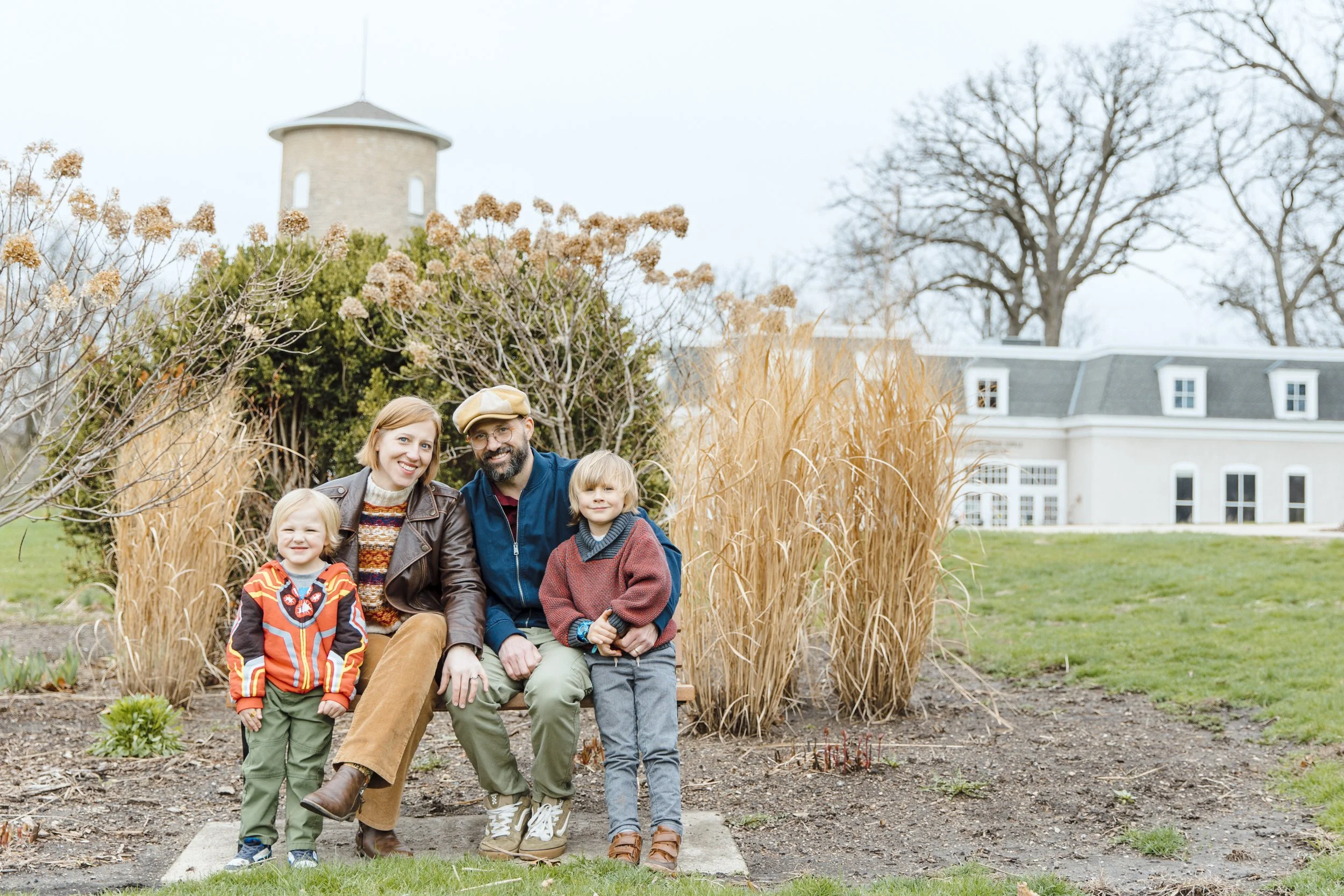 A family of five smiling for a photo outdoors, seated on a bench amid fall foliage with a large house and leafless trees in the background.