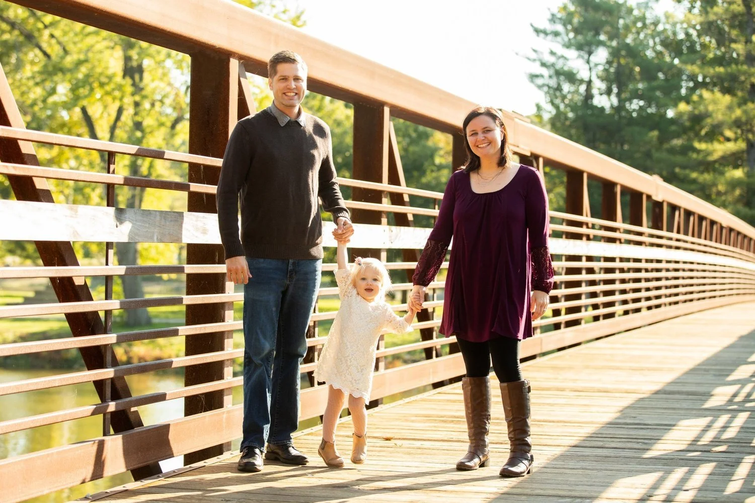 A family of three holding hands while walking on a wooden bridge outdoors during daytime. The man is wearing a dark sweater and jeans, the woman is dressed in a purple dress with black leggings and boots, and the young girl is in a light-colored dress.