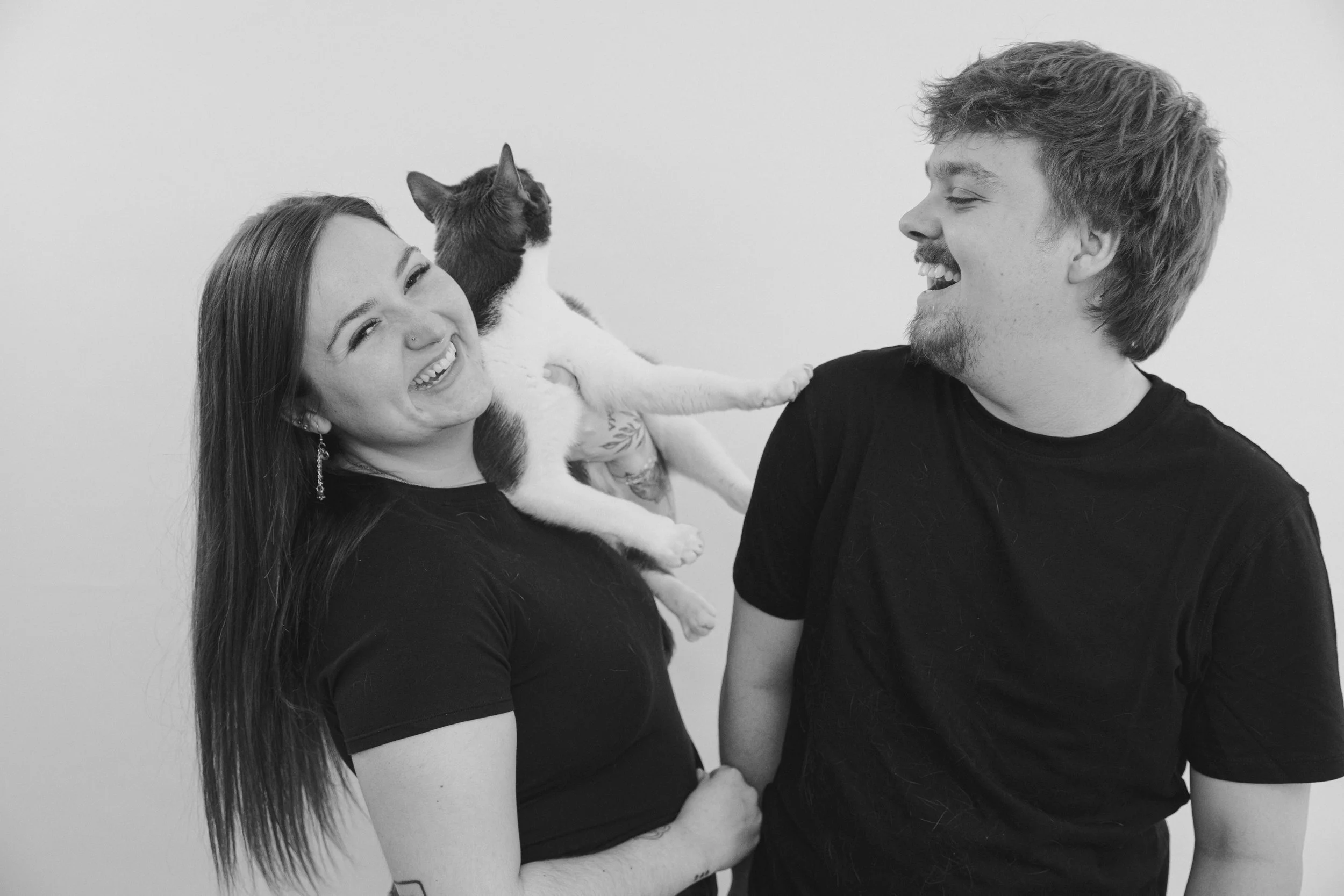 A woman and a man smiling at each other, while the woman holds a cat with white and dark fur. The woman has long hair and a nose piercing, and the man has tousled hair and facial hair. The background is plain.