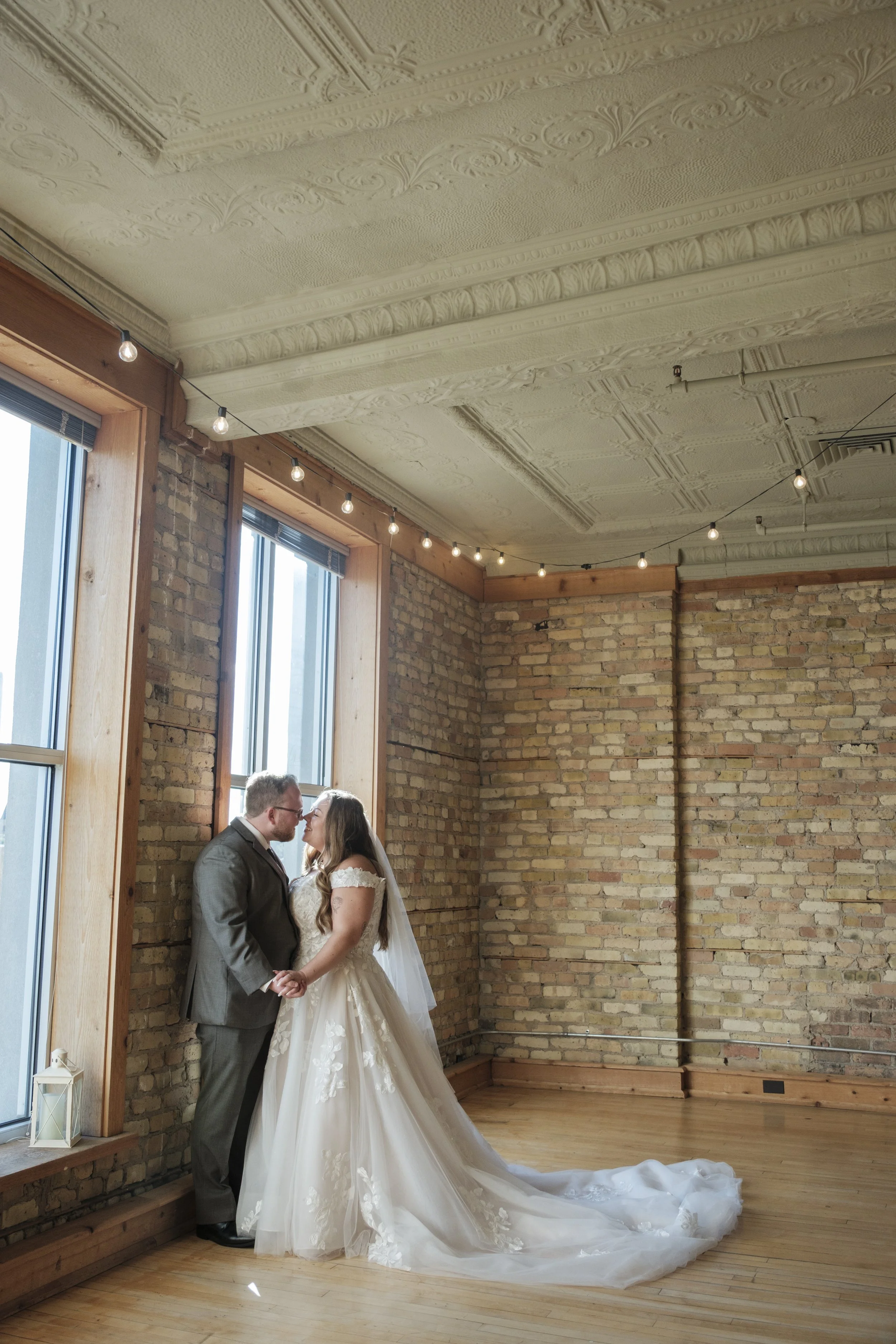 A bride and groom standing close, holding hands, and leaning in for a kiss inside a rustic brick-walled room with string lights and large windows.