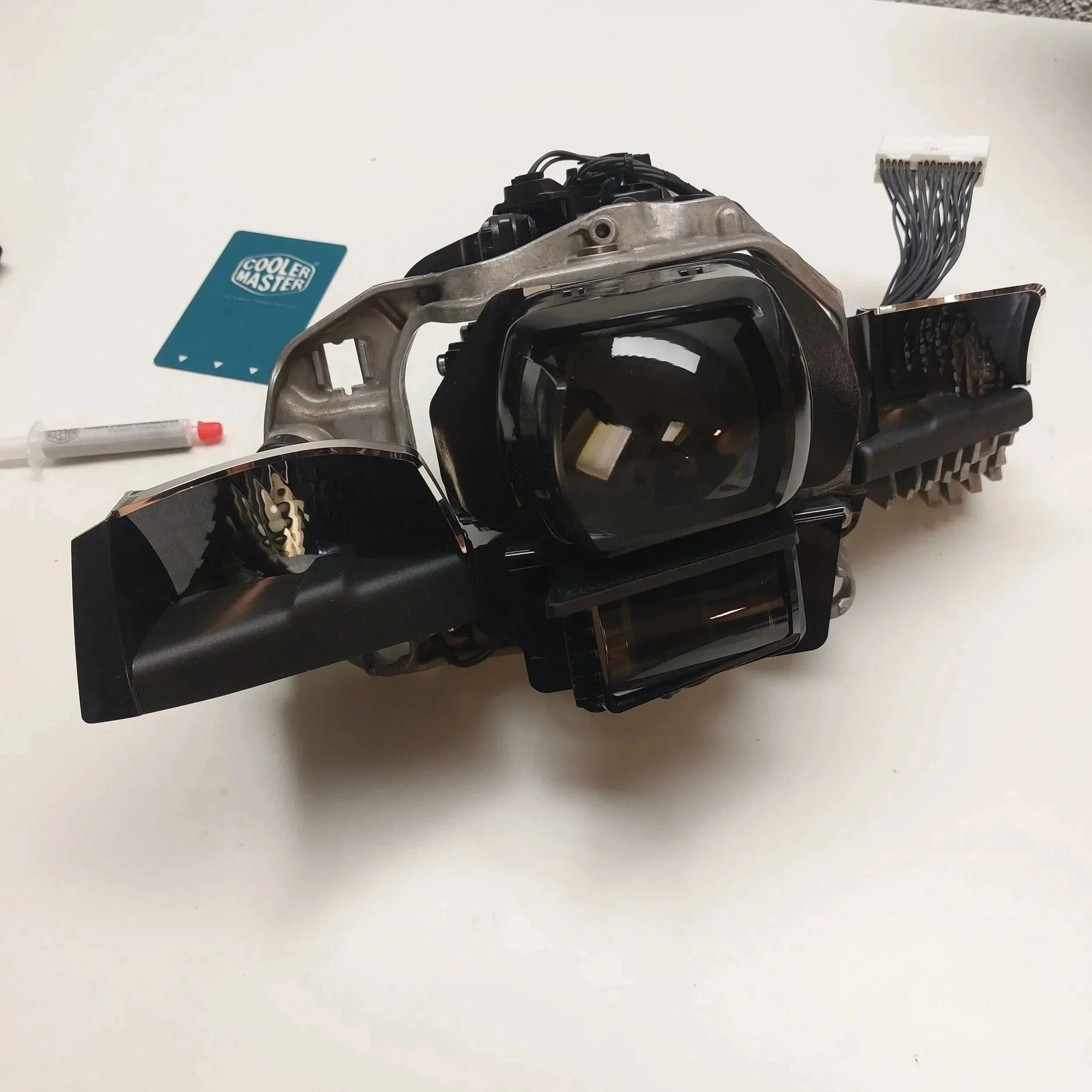 Automotive headlight assembly with wiring harness on a white surface, Cooler Master sticker, syringe with red cap nearby.