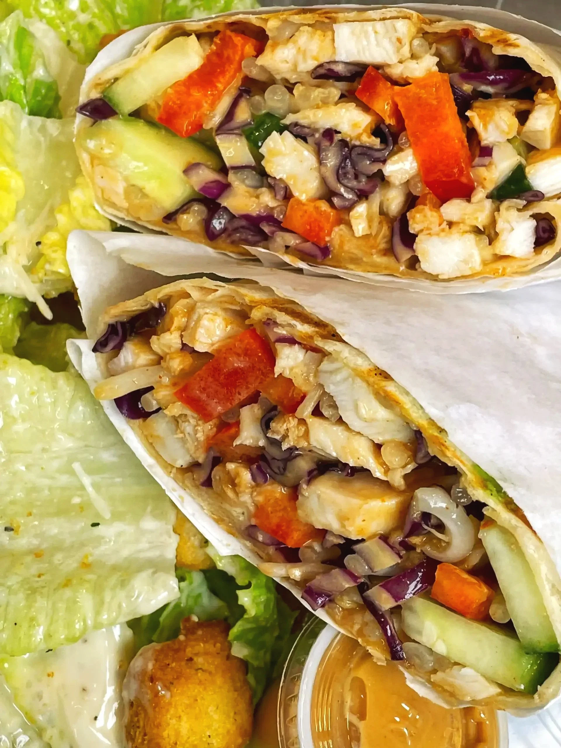 Two chicken and vegetable wraps cut in half, filled with chicken, red bell peppers, cucumbers, grapes, and purple cabbage, served with side salad and dipping sauce.