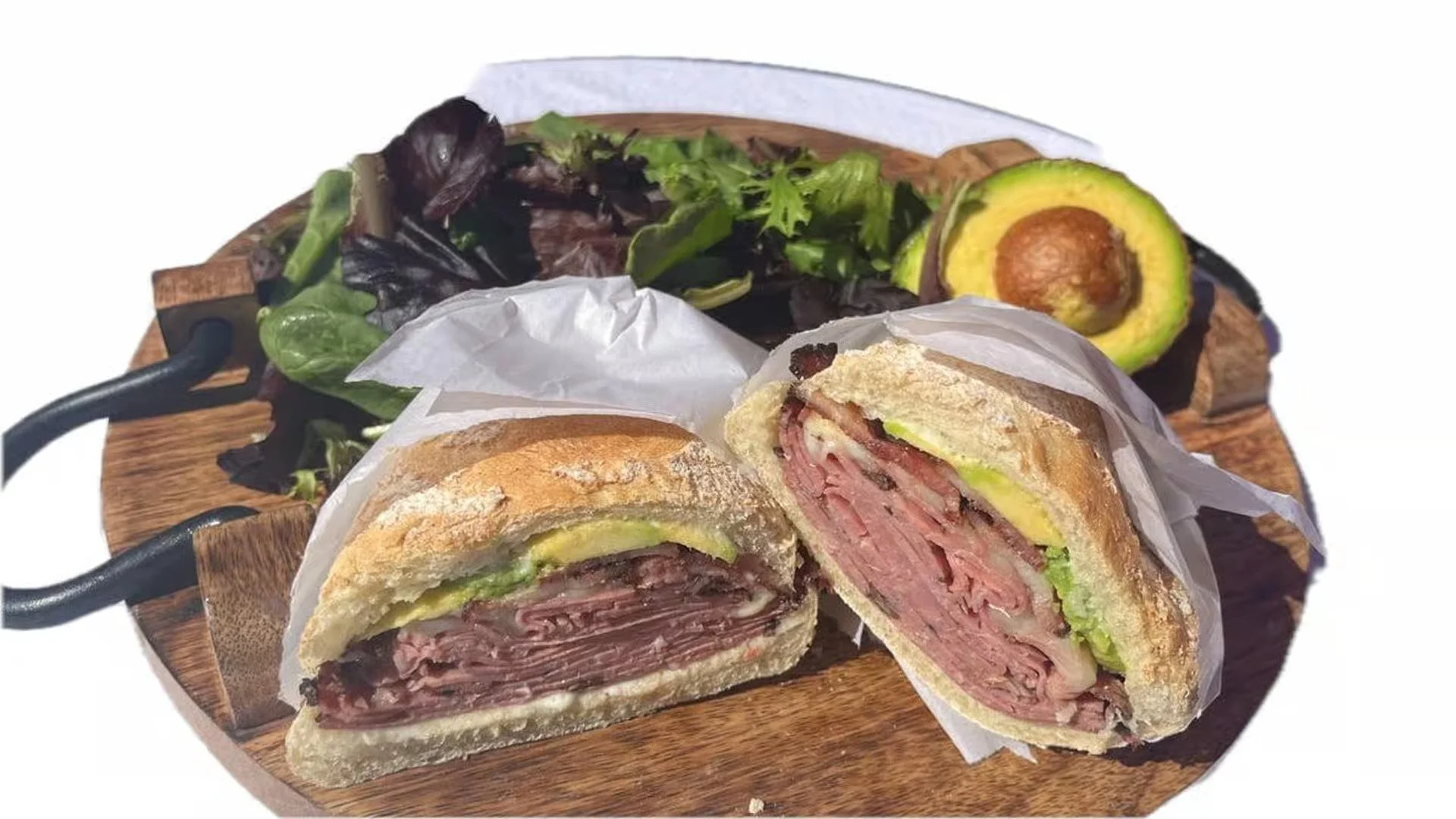 Sandwich with layers of thinly sliced cured meat, cheese, and avocado on a toasted roll, served on a wooden platter with leafy greens and an avocado half in the background.