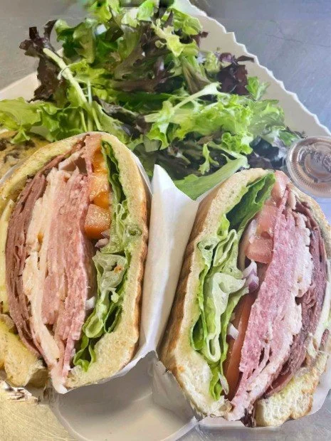 A sandwich with layers of ham, lettuce, tomato, and cheese, cut in half, served with a side of mixed greens salad.