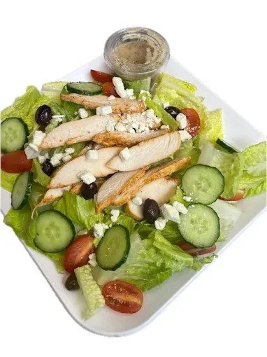 Caesar salad with grilled chicken, cherry tomatoes, cucumbers, black olives, crumbled cheese, and a small container of dressing on a white plate.