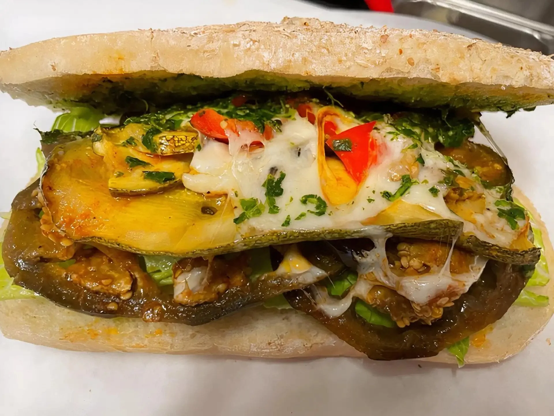 Close-up of a sandwich with roasted eggplant, melted cheese, chopped herbs, and sliced red chili peppers between two pieces of bread.