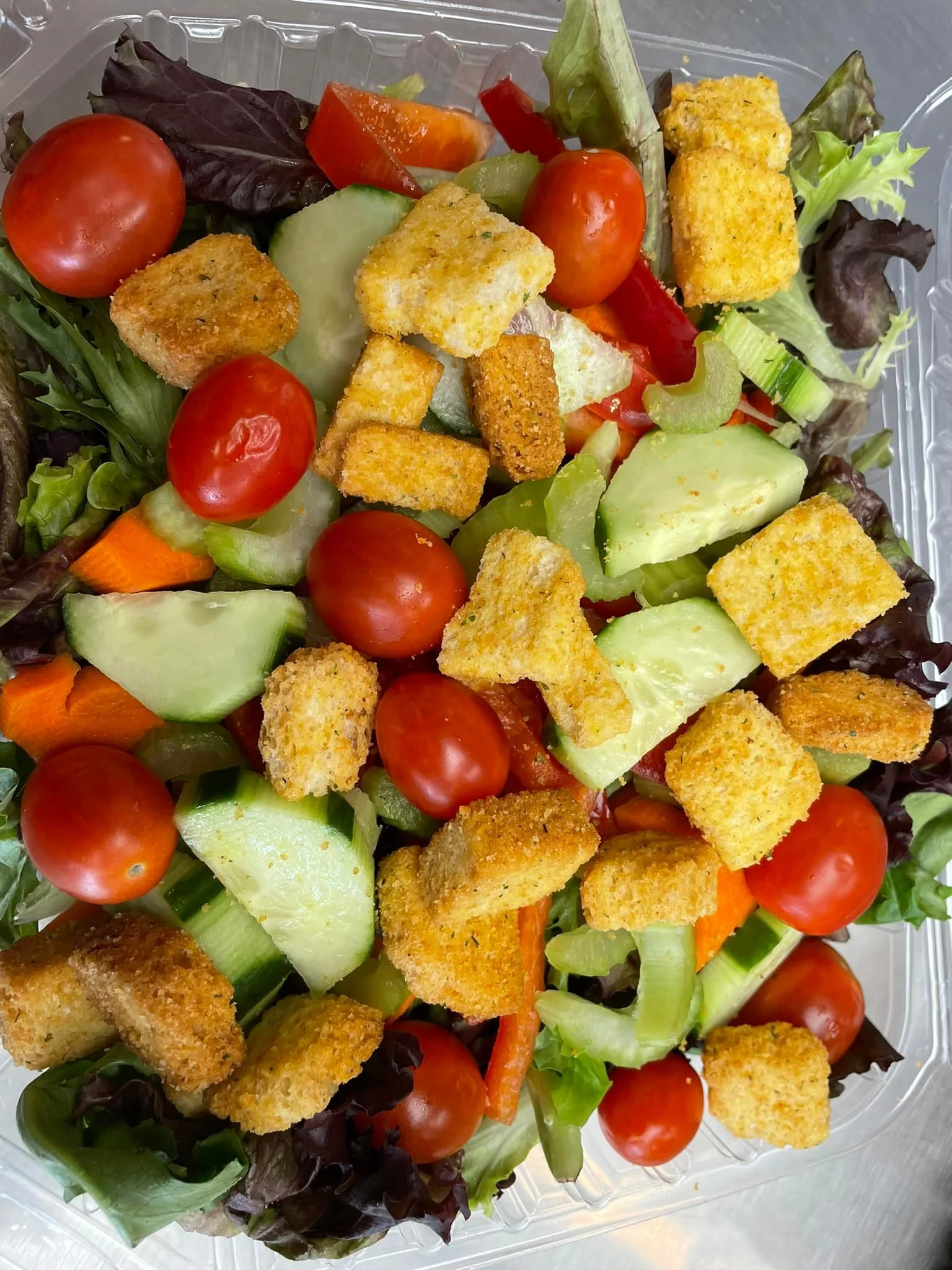 Mixed salad with cherry tomatoes, cucumbers, carrots, celery, red bell peppers, and croutons in a clear plastic container.