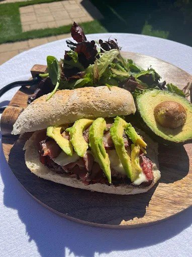 A wooden plate with a hot dog topped with guacamole, bacon, and other toppings, alongside a seeded bread roll and a side salad of mixed greens, seen outdoors with sunlight.