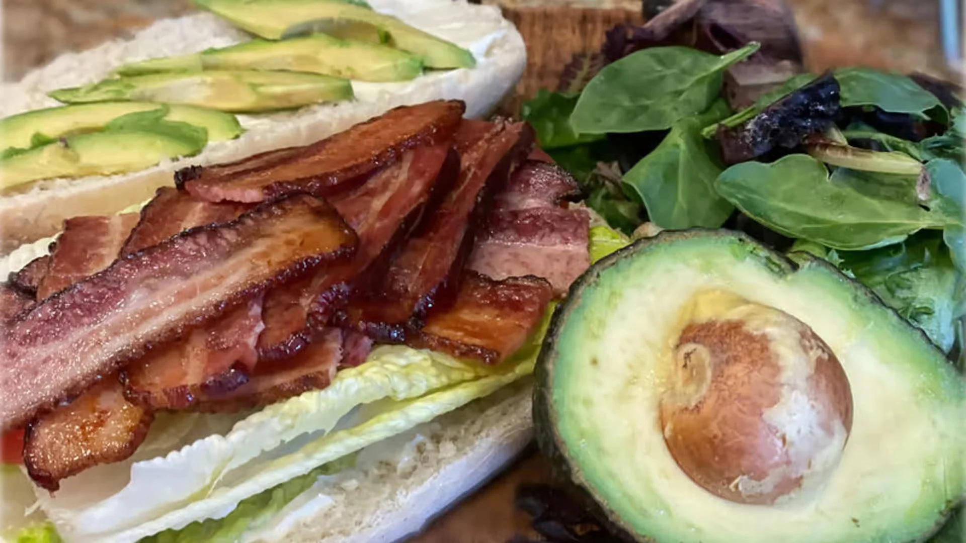 Close-up of a sandwich with bacon, avocado, and lettuce, served with a halved avocado on the side and fresh greens.