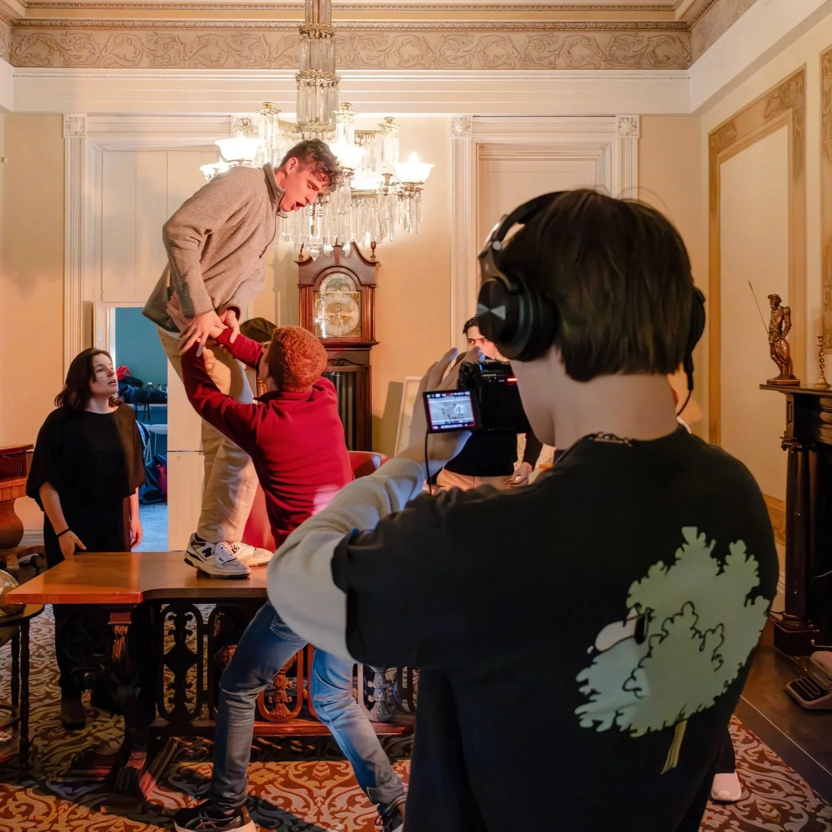 Film crew recording a scene of young actors performing in a living room with ornate decor and a chandelier.