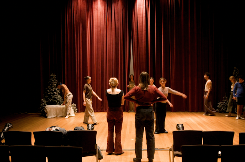 People rehearsing in front of a stage with red curtains, some with arms extended, and a few seated chairs in front.