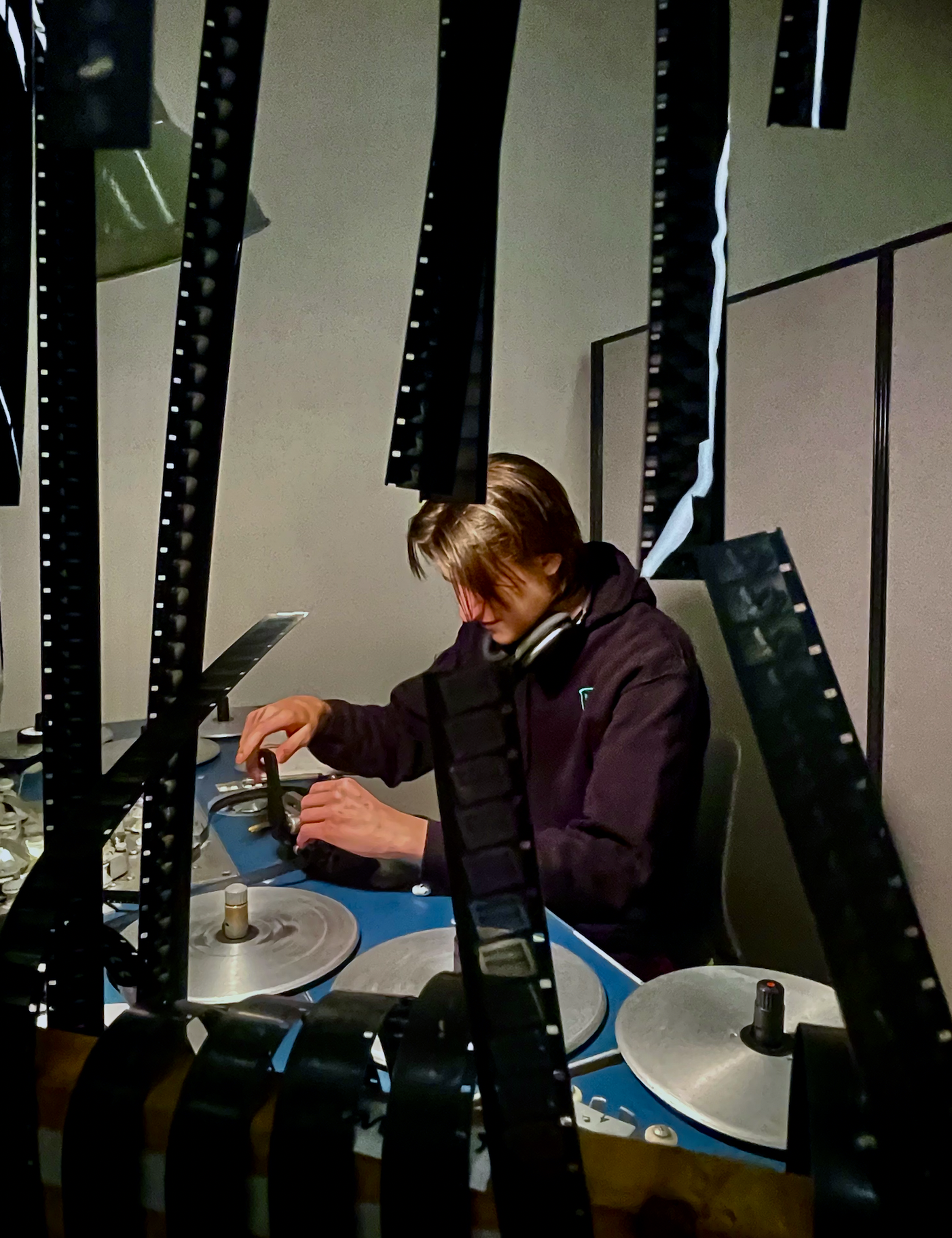 A person with shoulder-length brown hair working at a desk with multiple film strips and a computer. The person is wearing a dark hoodie and has headphones around their neck.