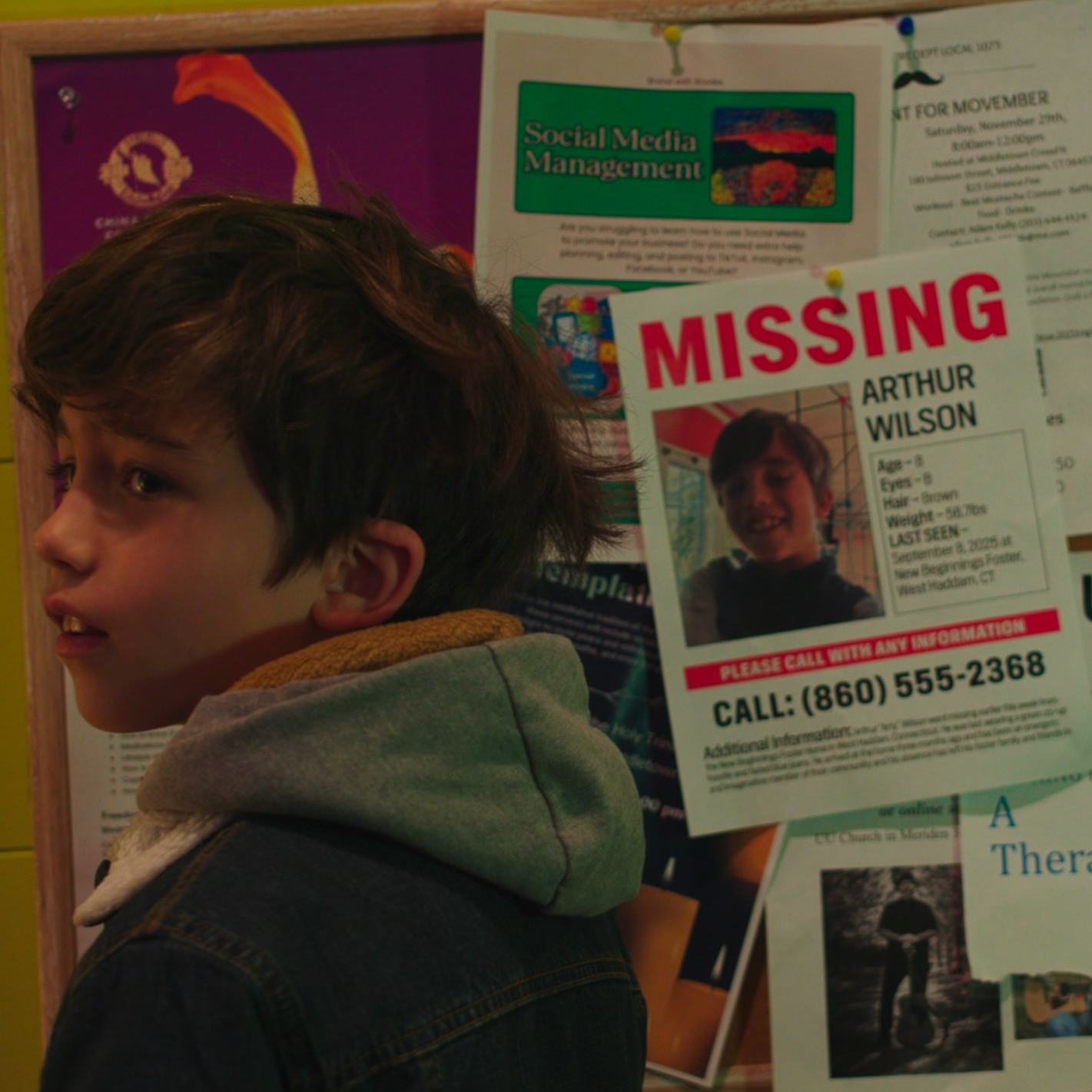 A young boy with brown hair and a gray hoodie looking sideways in front of a missing person flyer with the name Arthur Wilson.