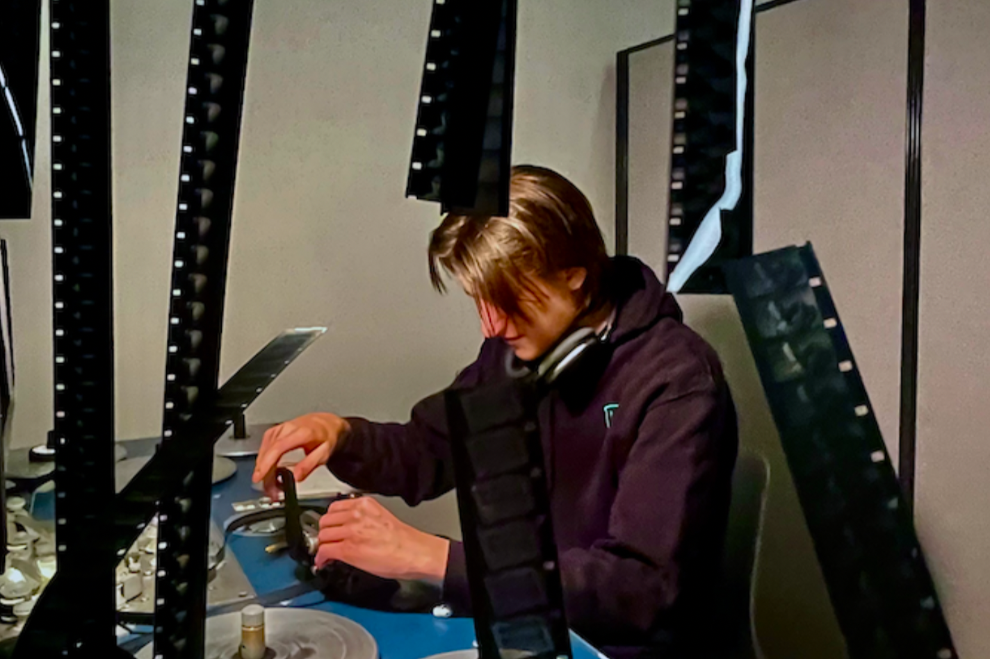 A person with shoulder-length brown hair working at a desk with multiple film strips and a computer. The person is wearing a dark hoodie and has headphones around their neck.