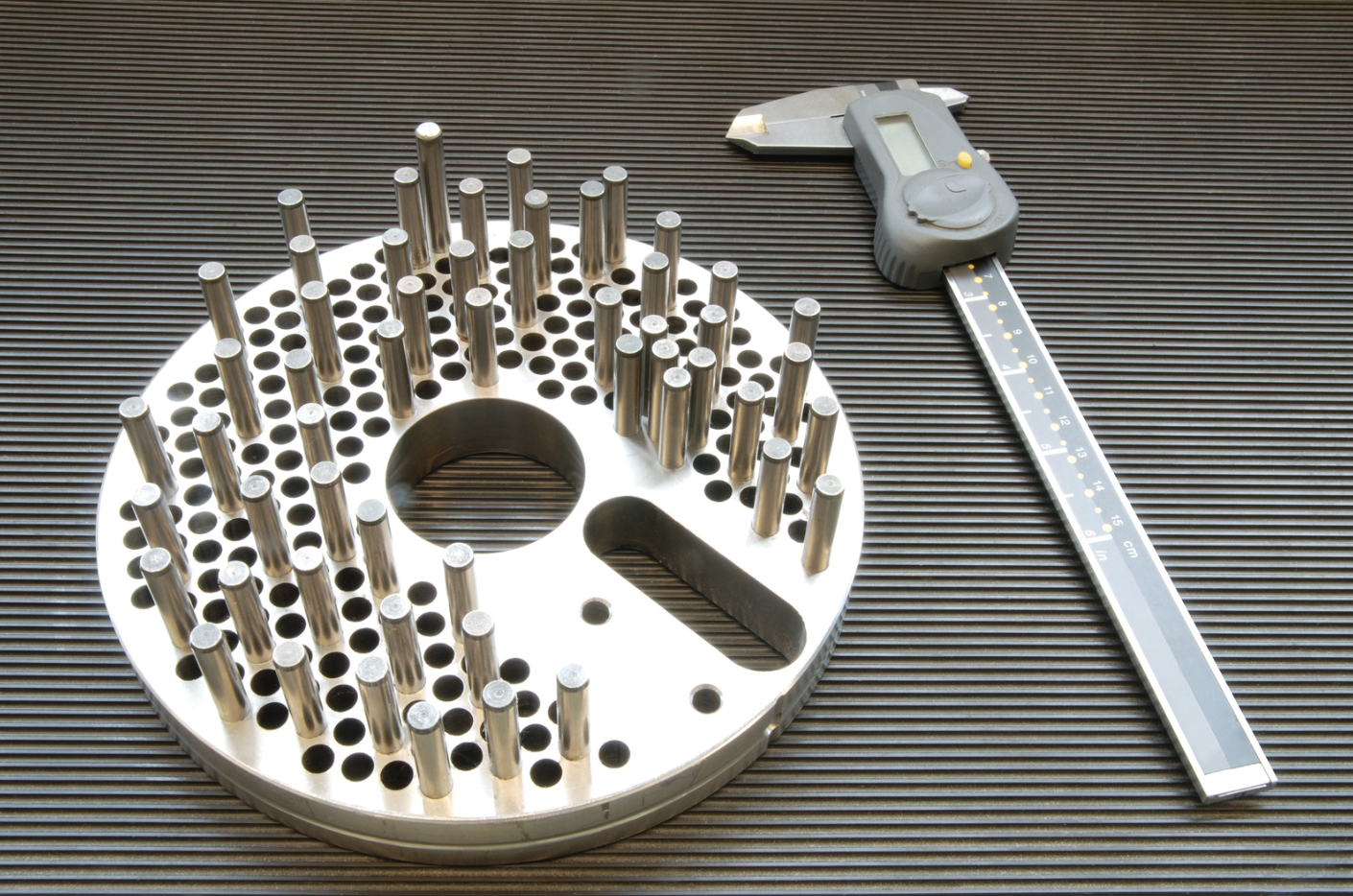 A circular metal component with many evenly spaced cylindrical pins, a hole in the center, and a cutout segment, placed on a black ribbed surface. A measuring tape is positioned to the right of the component.