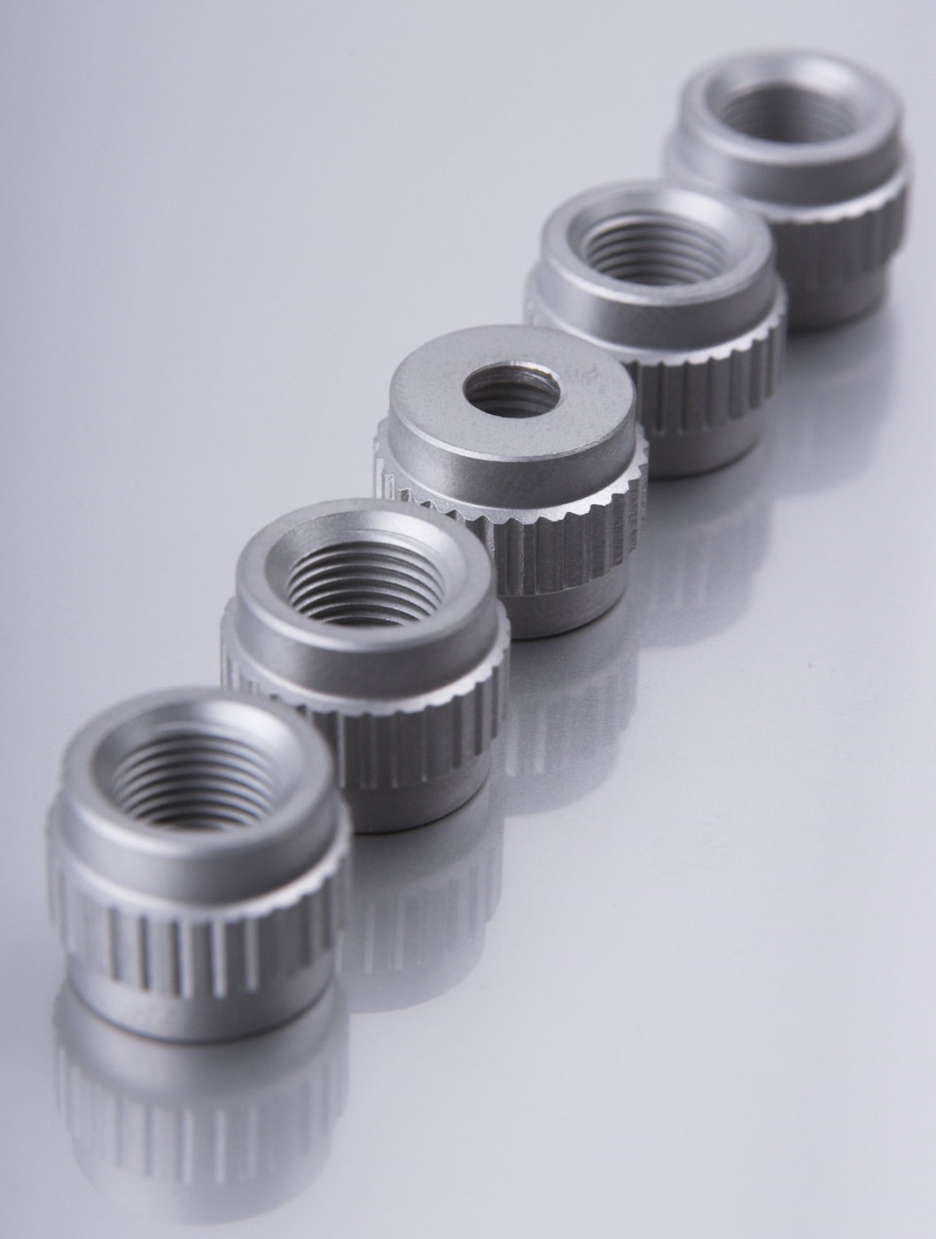 A row of six metal threaded components with knurled edges, placed on a reflective surface.