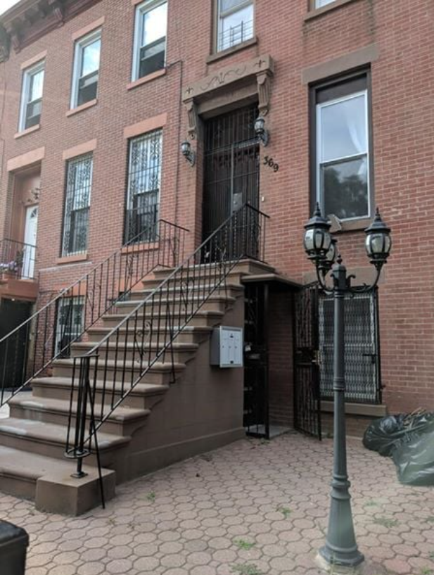 A brick apartment building with a staircase leading up to the front door, which has black wrought iron gates. The building has several windows, some with metal bars. There is a modern lamp post and a trash bag on the sidewalk.