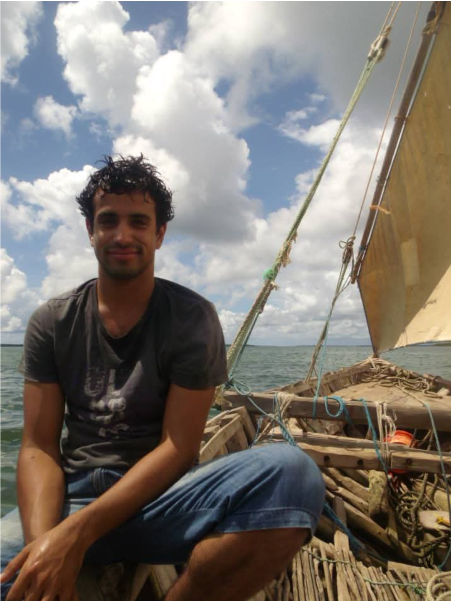 A young man with dark curly hair and a beard, wearing a black T-shirt and blue shorts, sitting on a wooden boat with sails and ropes, during the daytime with a cloudy sky and water in the background.