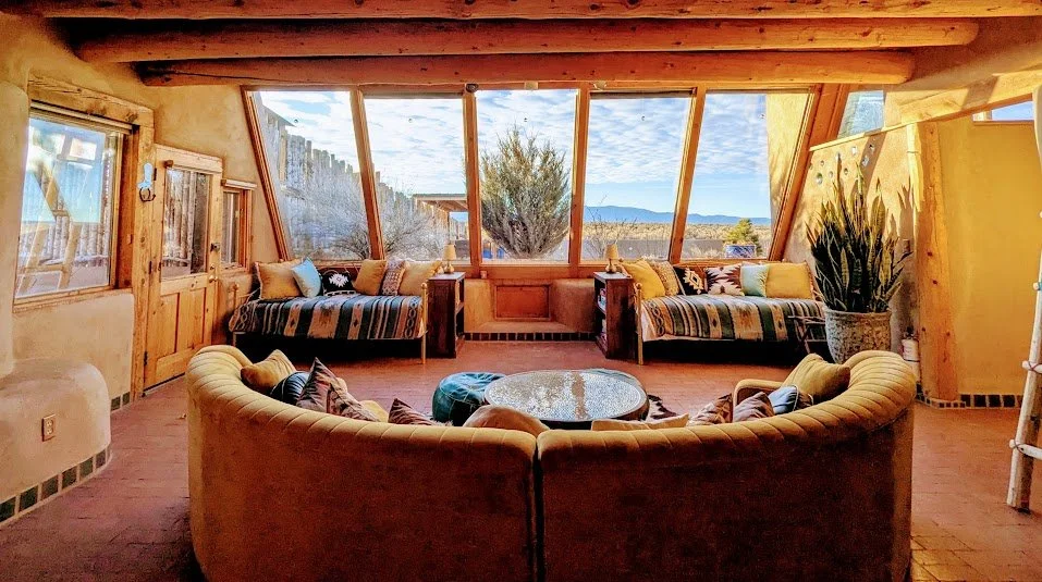 A cozy living room with large angled windows showcasing a desert landscape with bushes and mountains in the background. The room has wooden beams, a rounded yellow sofa with pillows, two striped sofas with pillows, a round coffee table, and a tall potted plant.