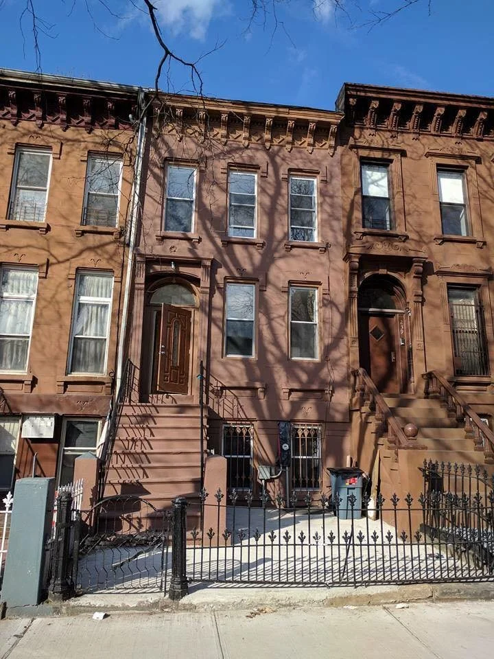 Three-story brownstone row houses with decorative window frames, front steps, small yards enclosed by black iron fences, and a blue sky with a tree shadow.