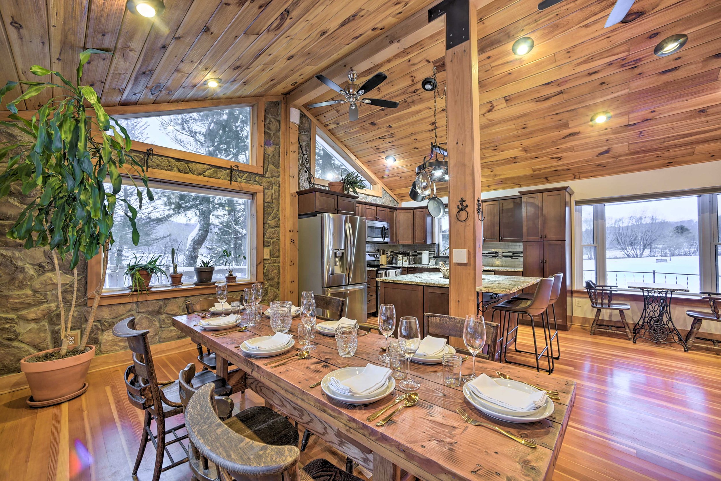 A rustic kitchen and dining area with wooden ceiling, stone wall, large windows showing snow outside, a wooden dining table set with white plates, glasses, and gold utensils, a potted plant, kitchen appliances, and barstools.