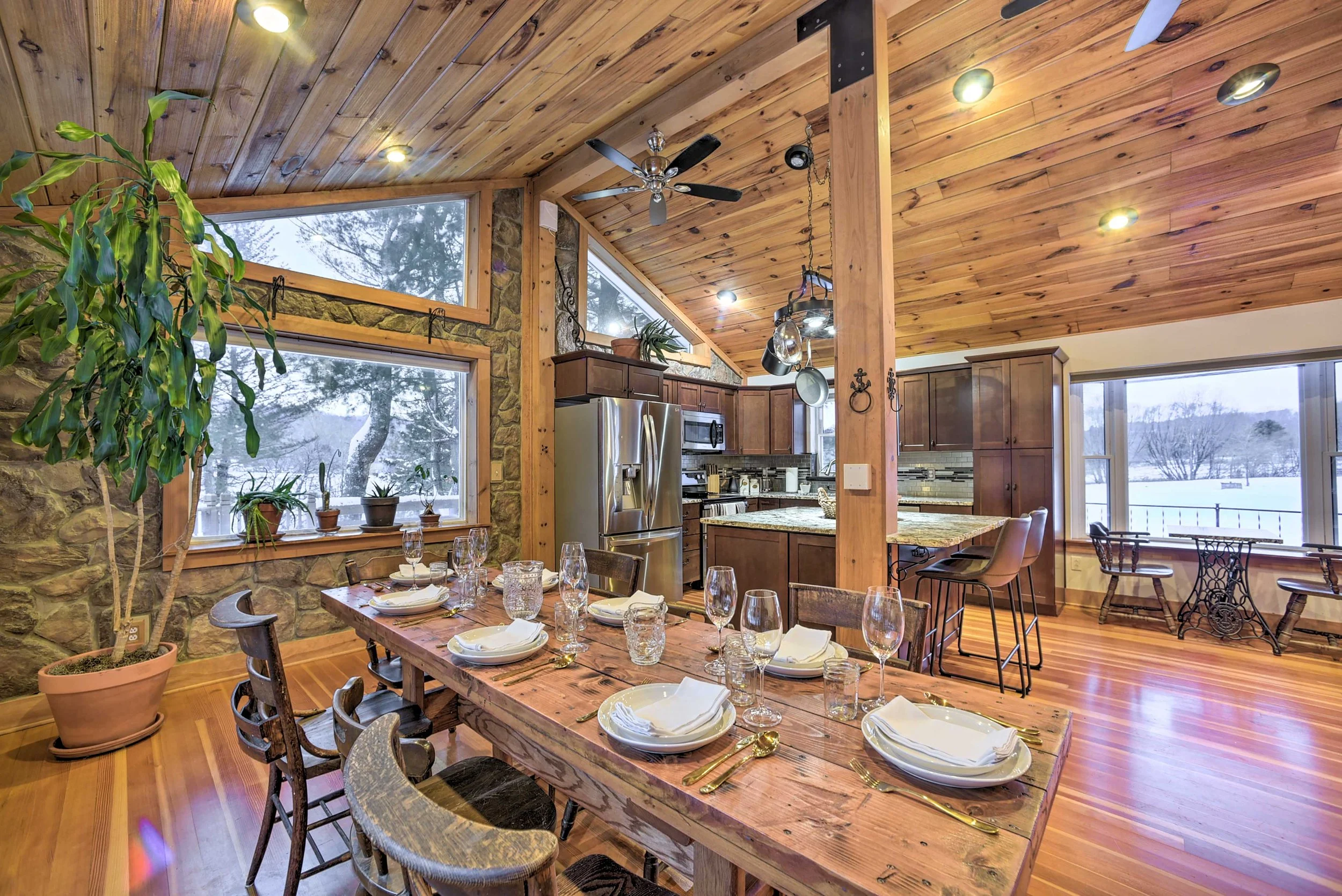 A rustic kitchen and dining area with wood-paneled ceiling and floors, stone walls, large front window and larger rear window showing snowy landscape outside, round ceiling fans, hanging pots, cabinet kitchen with granite countertops, stainless steel fridge, and a dining table set with plates, glasses, and cutlery.