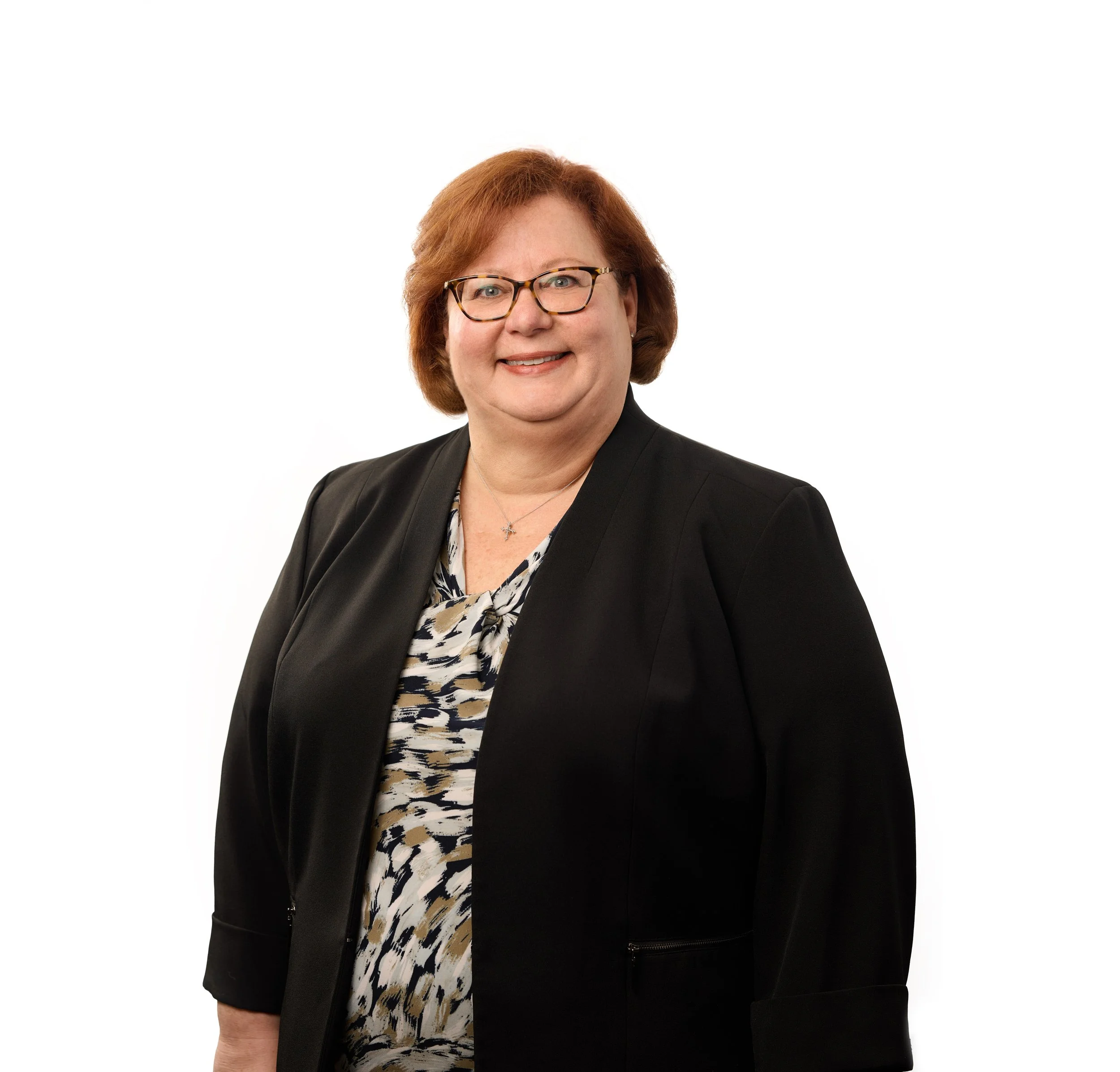 A woman with short red hair and glasses smiling, wearing a black blazer and a printed blouse, against a plain white background.