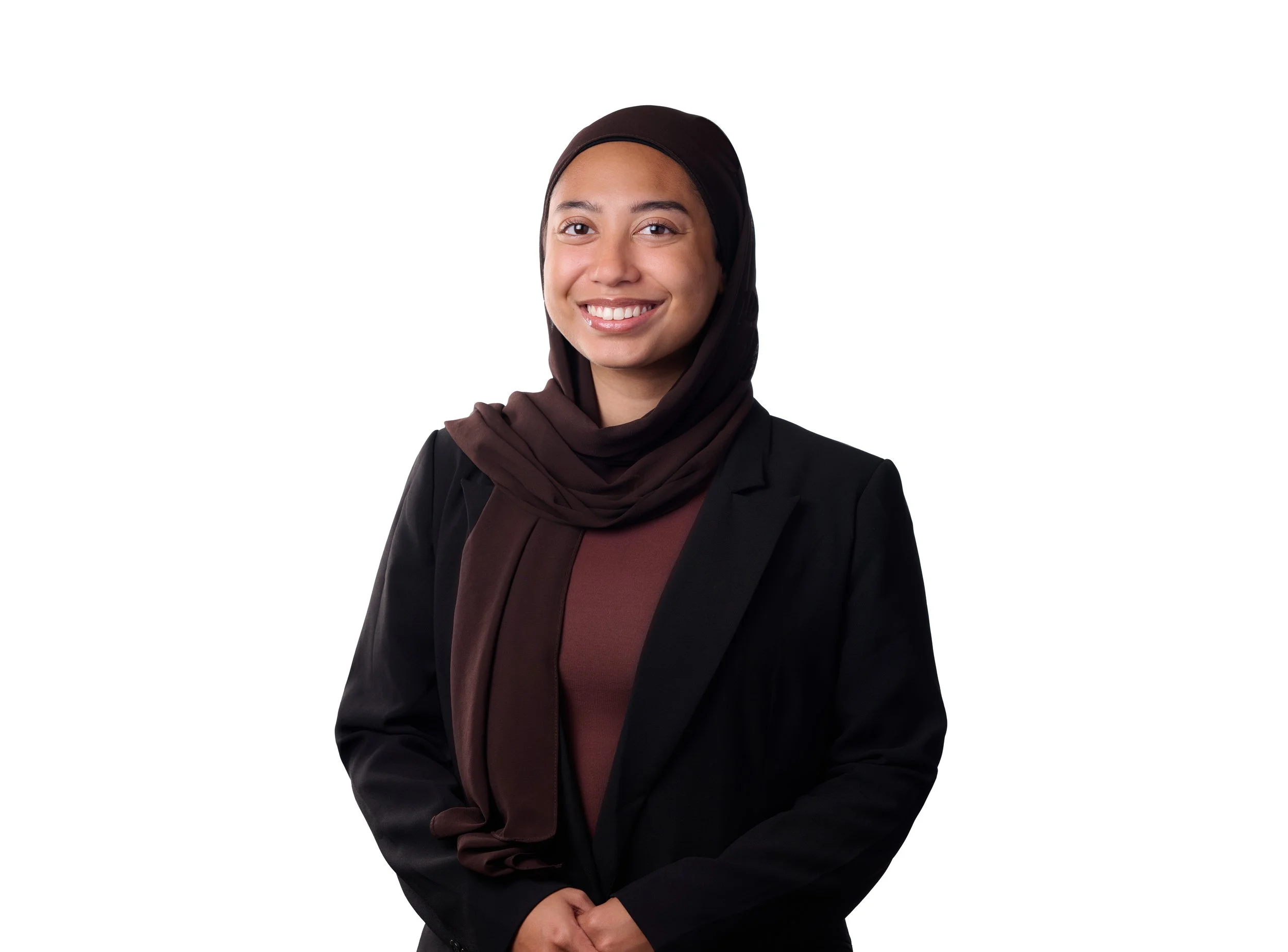 Portrait of a young woman wearing a black hijab and blazer, smiling, against a plain white background.