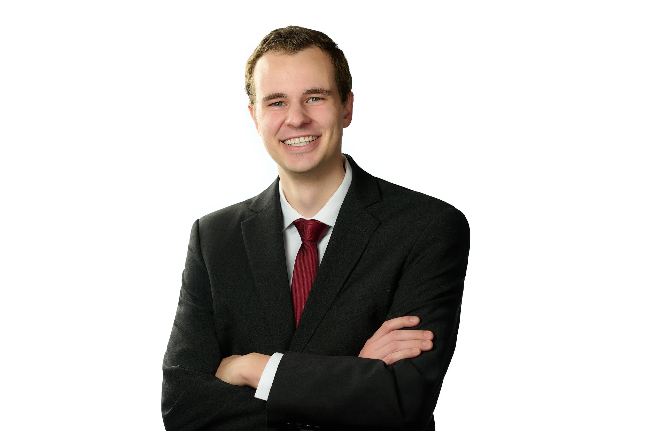 A young man in a black suit, white shirt, and red tie smiling with arms crossed against a plain white background.