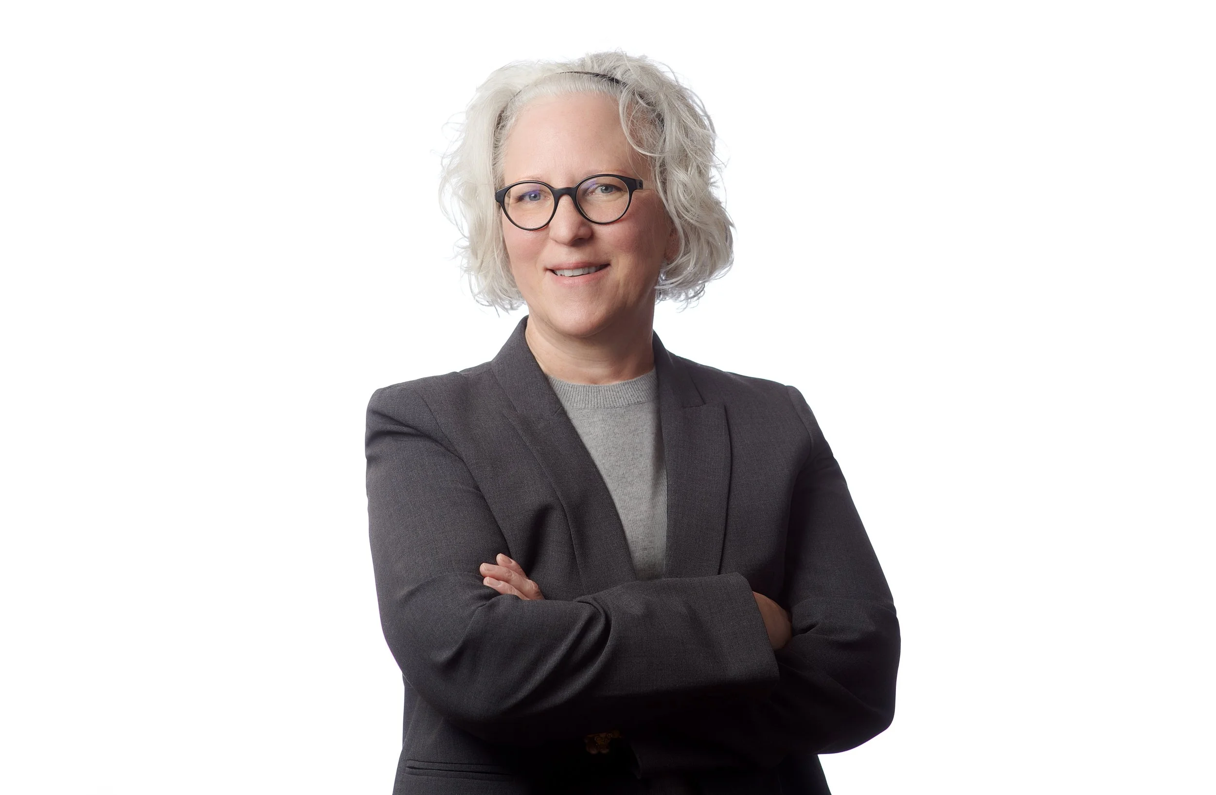 A confident middle-aged woman with curly gray hair and black glasses, wearing a gray sweater and a black blazer, standing with her arms crossed against a plain white background.