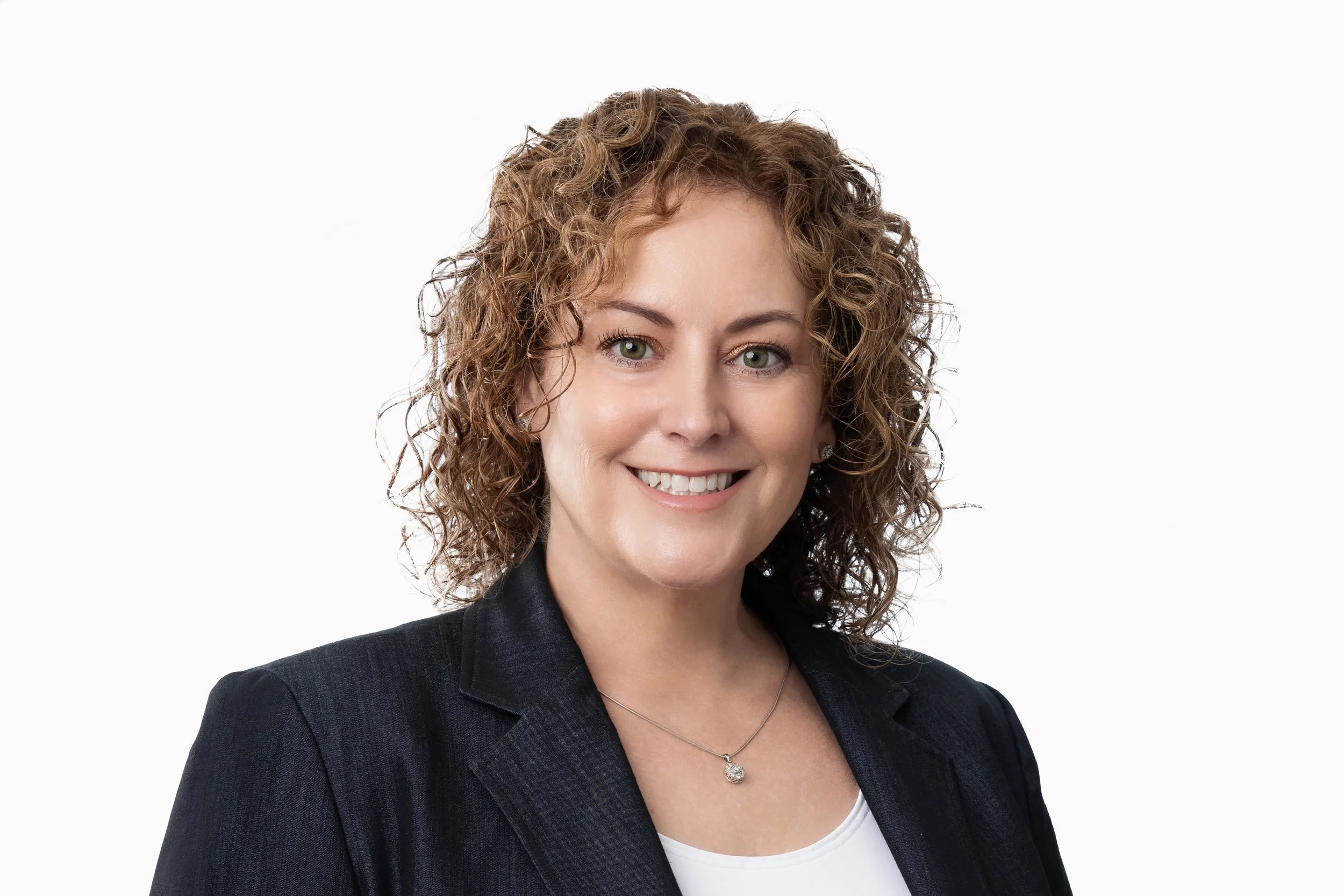 Headshot of a woman with curly brown hair, green eyes, wearing a dark blazer and a necklace, smiling against a plain white background.