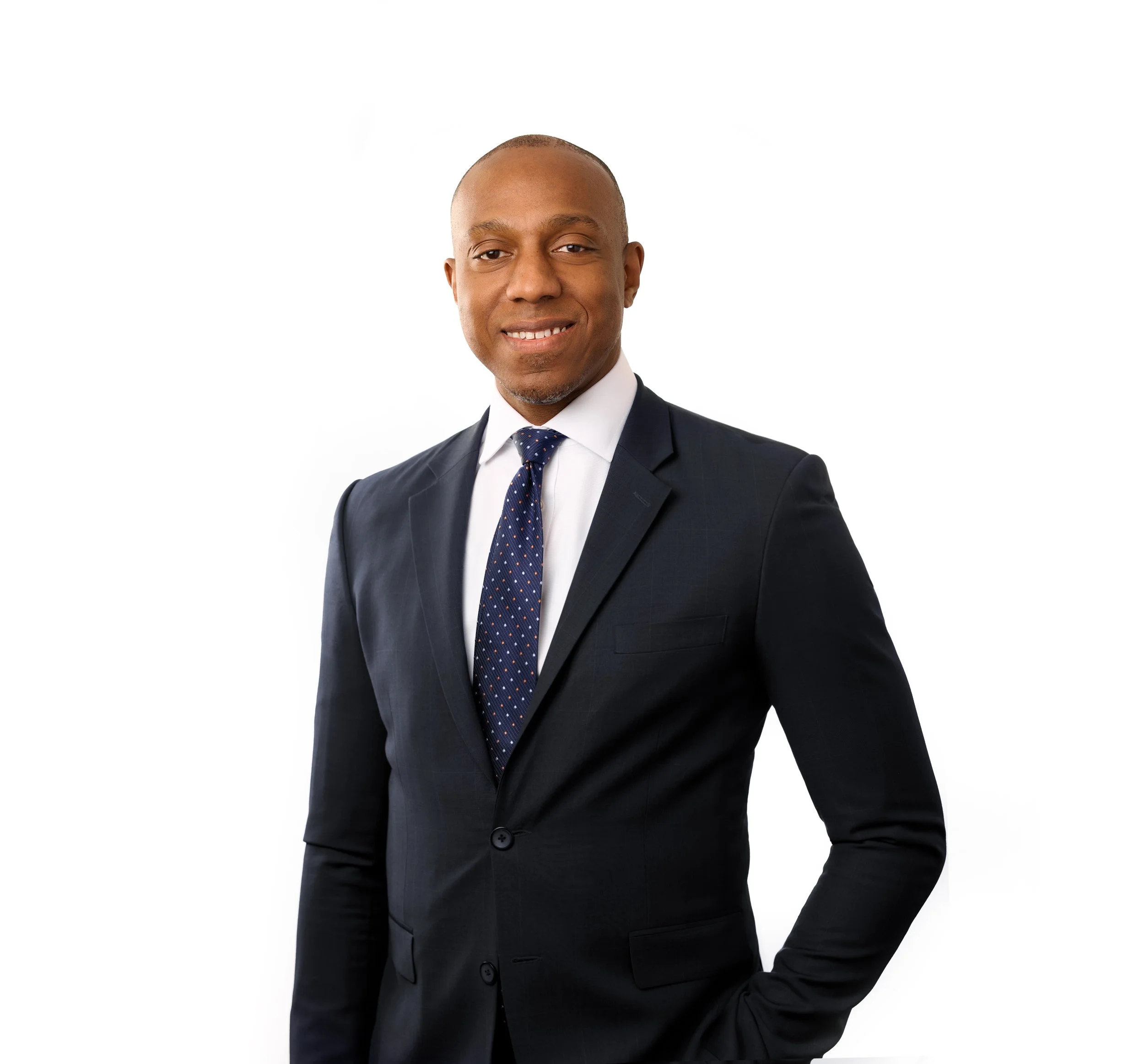 A man dressed in a dark suit with a white shirt and a dark blue patterned tie, smiling and posing with a hand on his hip, against a plain white background.