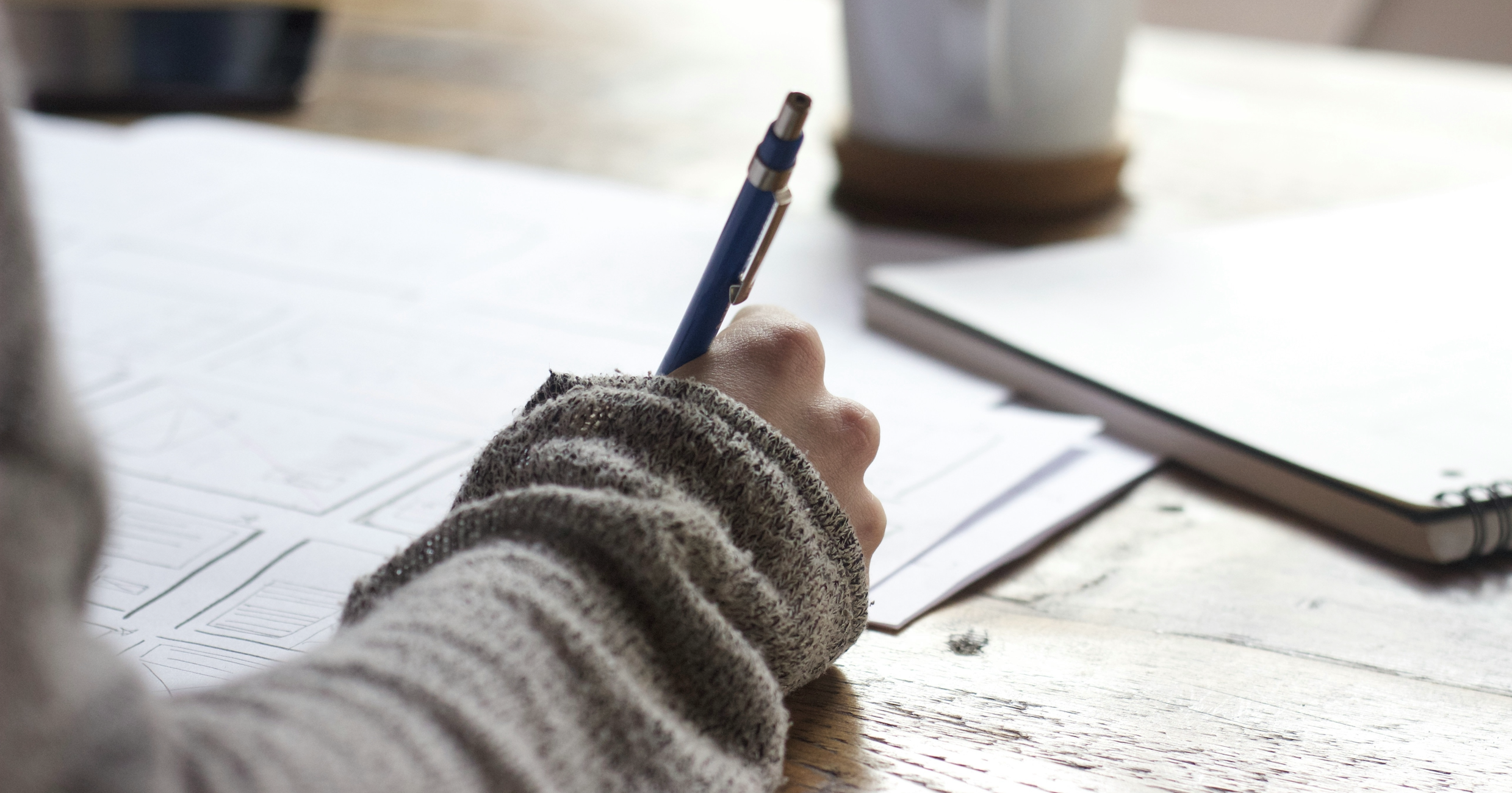 Une main qui tient un stylo, écrivant sur des feuilles de papier avec des croquis ou des plans, sur une table en bois.