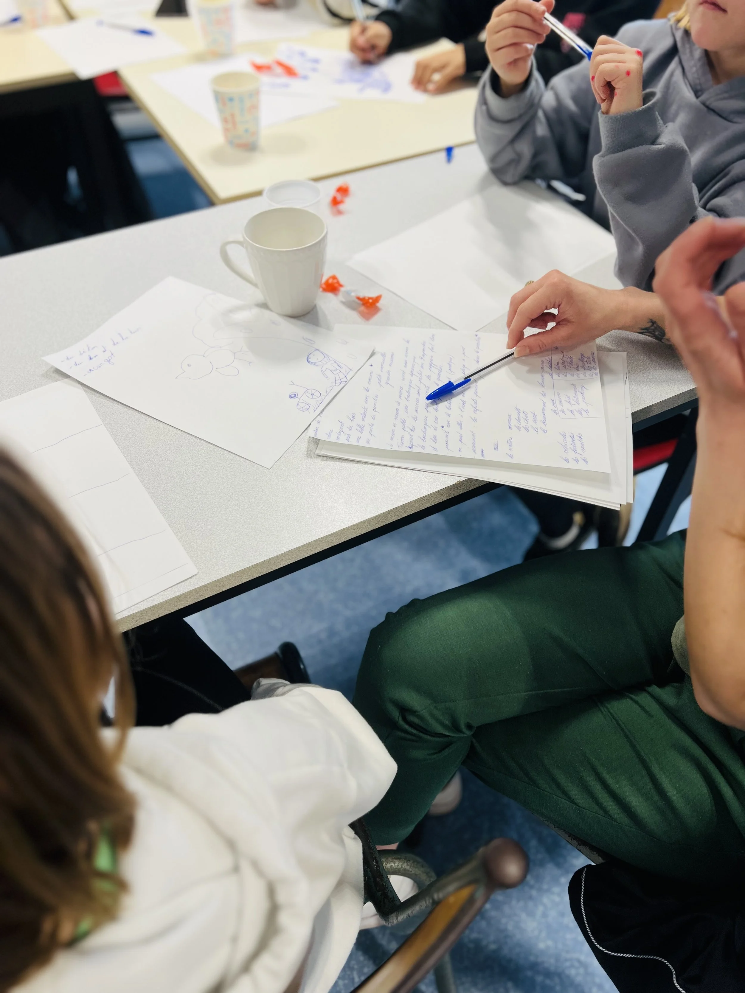 Groupe d'étudiants ou de personnes assises autour d'une table en train d'étudier ou de discuter, avec des feuilles, des stylos, des mugs et des bonbons sur la table.