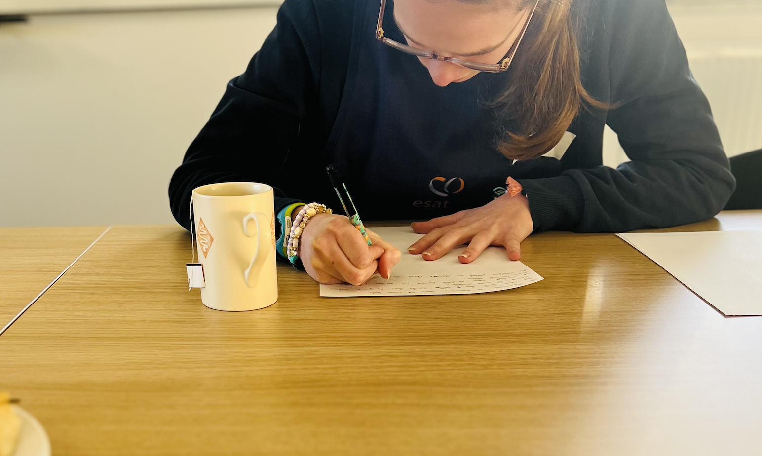 Une personne écrivant sur une feuille de papier posée sur une table en bois, avec une tasse beige à côté.