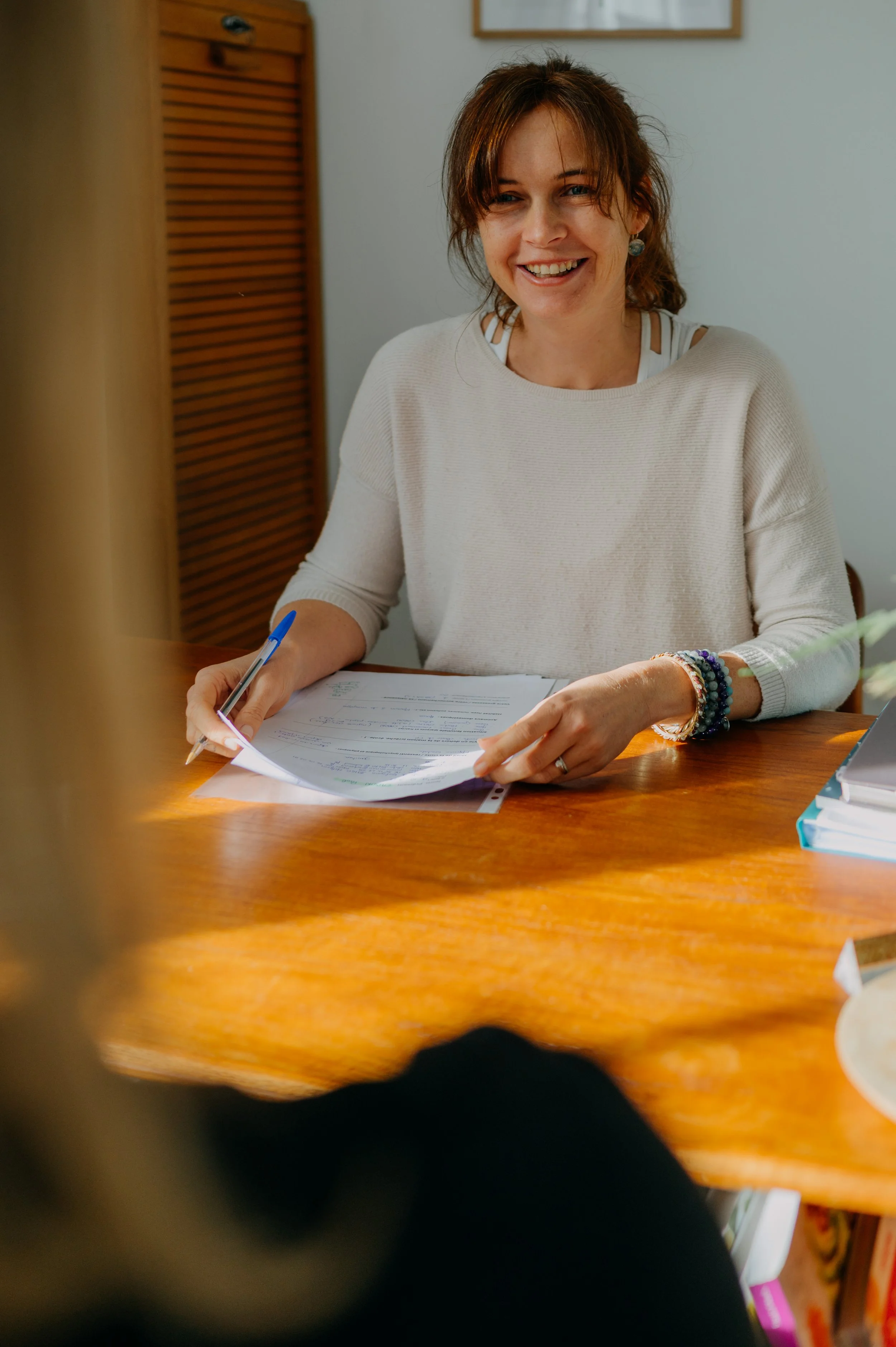 Julie Neveu, souriante assise à une table en bois, tenant des documents et un stylo, dans une pièce bien éclairée.