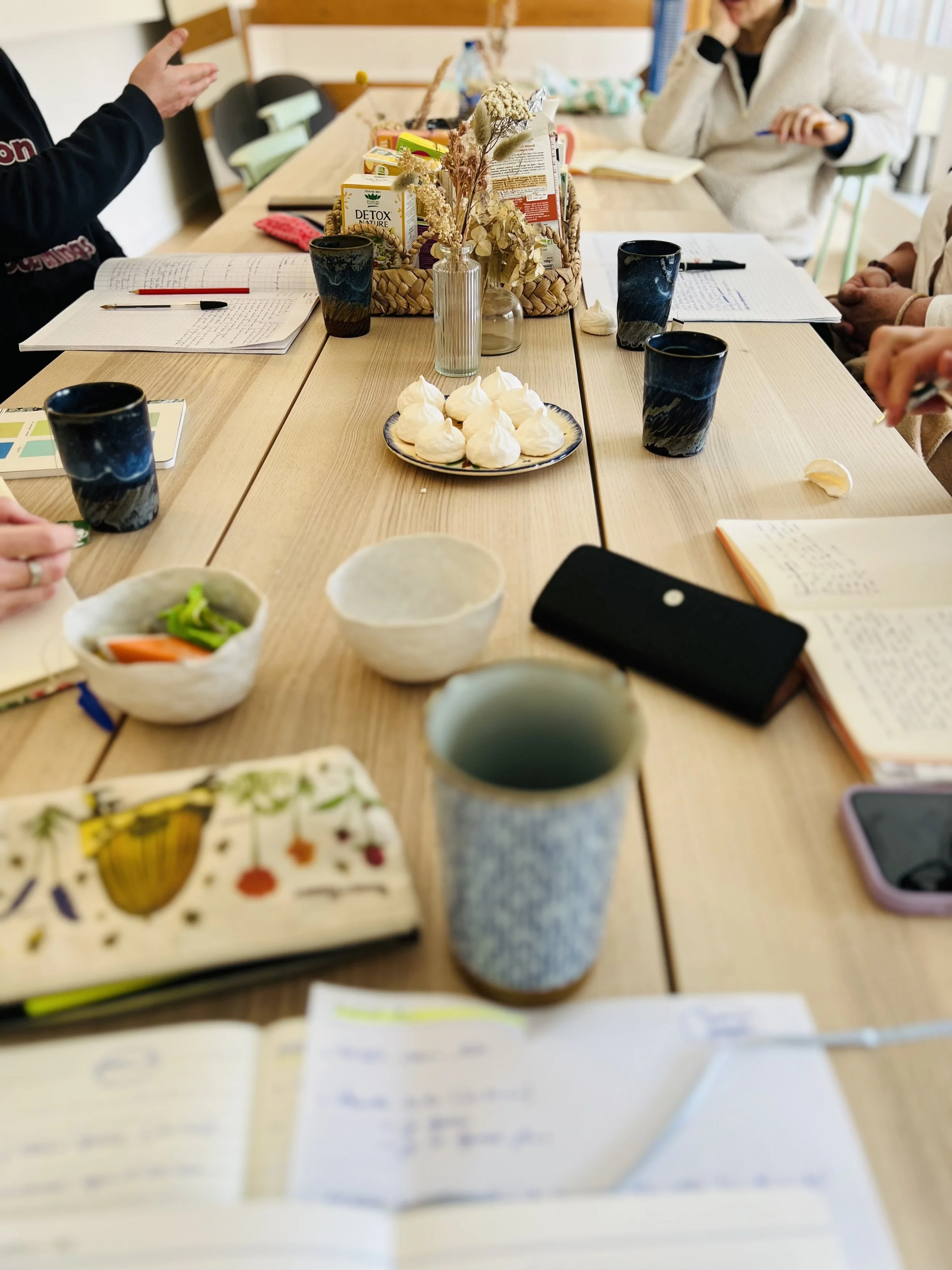 Une grande table conviviale avec du thé, café, des douceurs à grignoter, des carnets, des stylos, et des personnes discutant dans une ambiance chaleureuse.