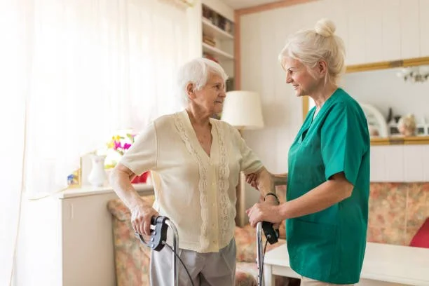 Nursing home caregiver helping an elderly woman with her walker in a bright, cozy room.