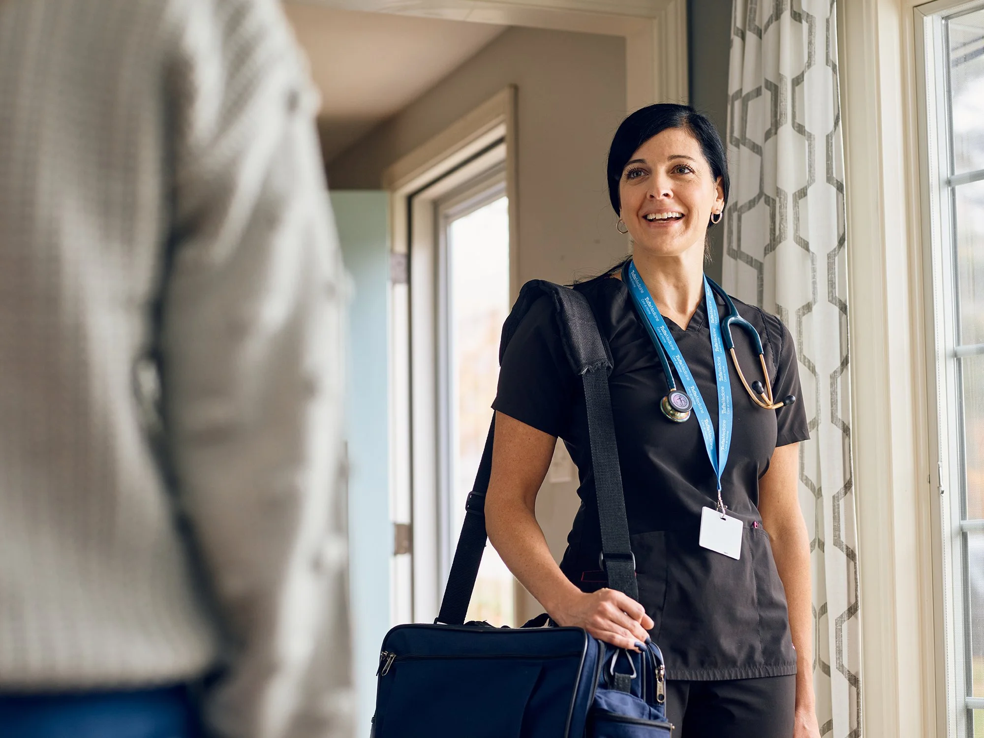 Female nurse or doctor in uniform smiling indoors by a window with curtains.