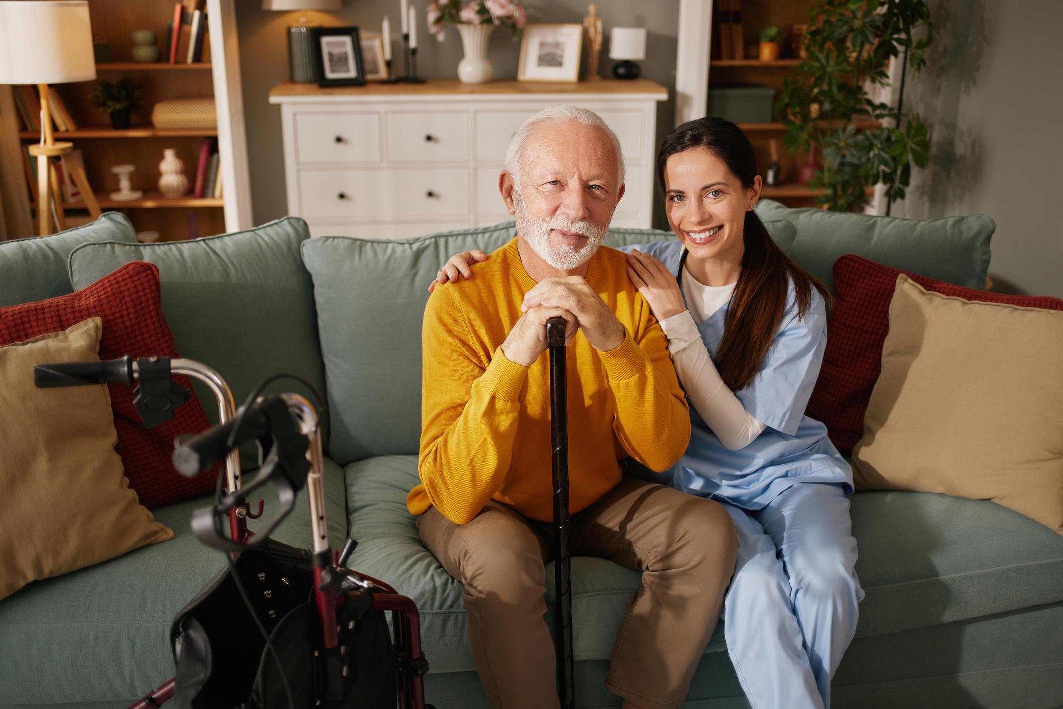 An elderly man with white hair and a beard sitting on a sofa with a cane, smiling, beside a young woman in medical scrubs, also smiling, in a cozy living room.